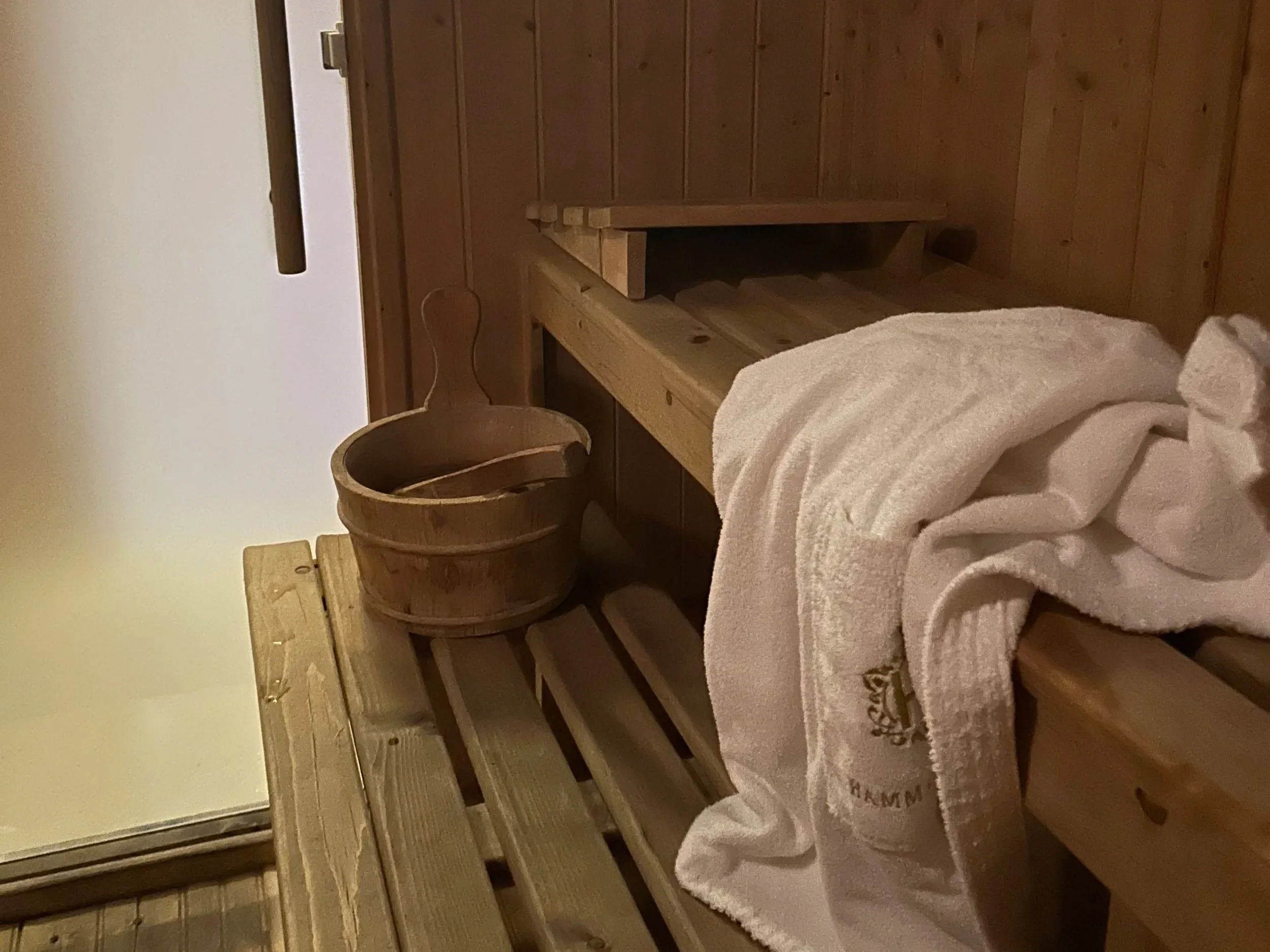 A rustic bedroom with wood-paneled walls and wooden furniture, including a bed with white towels and embroidered initials, a small side table, and a wooden ladder or support structure.