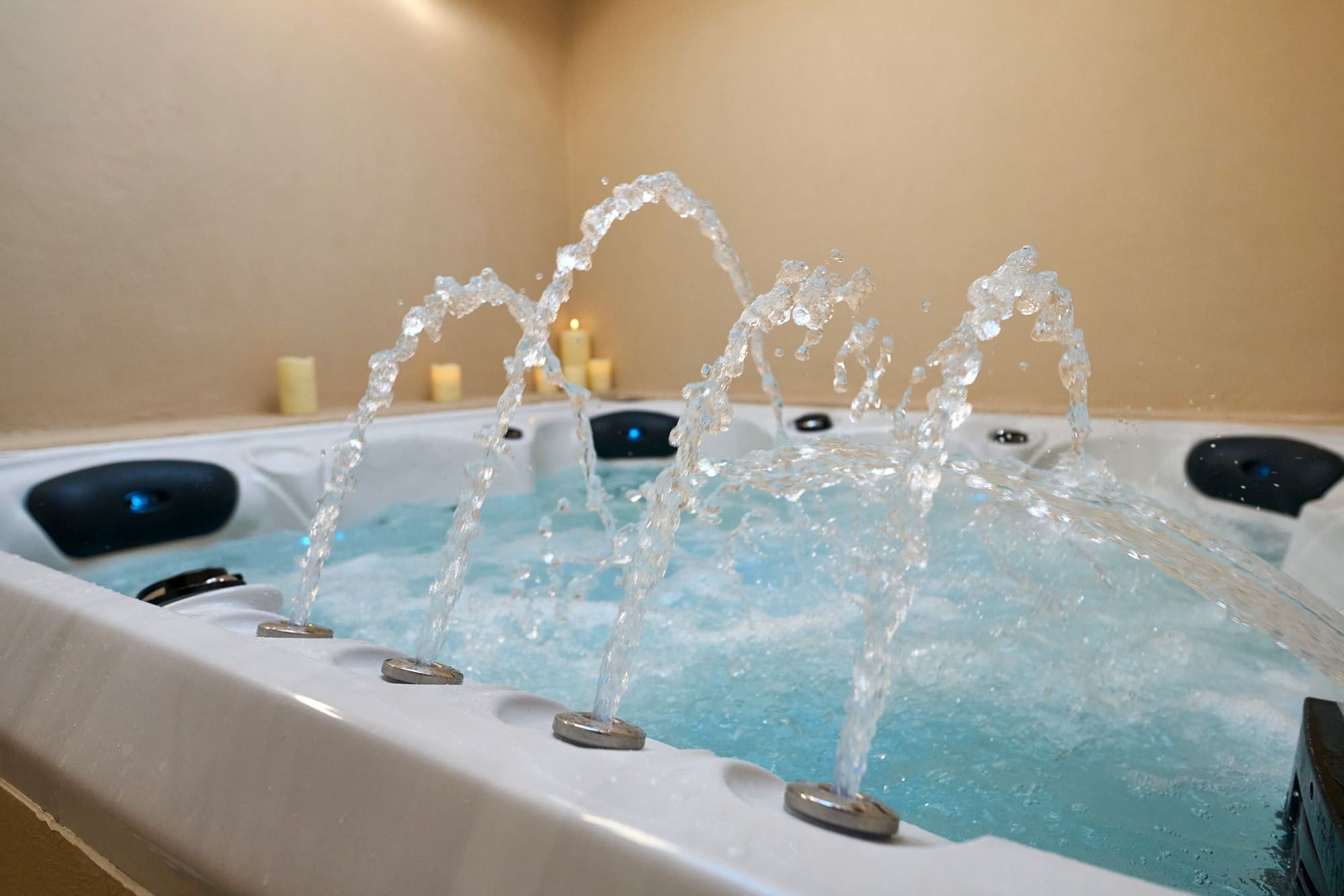 A hot tub with water jets spraying water inside, candle lights in the background, beige walls, and black headrests.