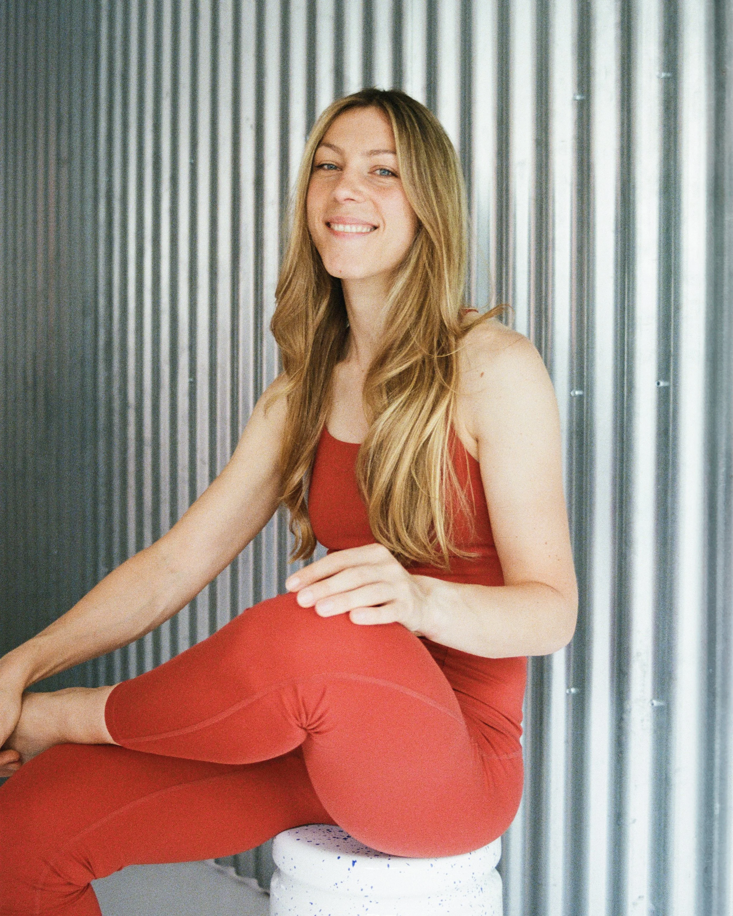 A woman with long blonde hair wearing a red sleeveless top and red leggings, sitting on a white stool, smiling, in front of a metallic corrugated wall.