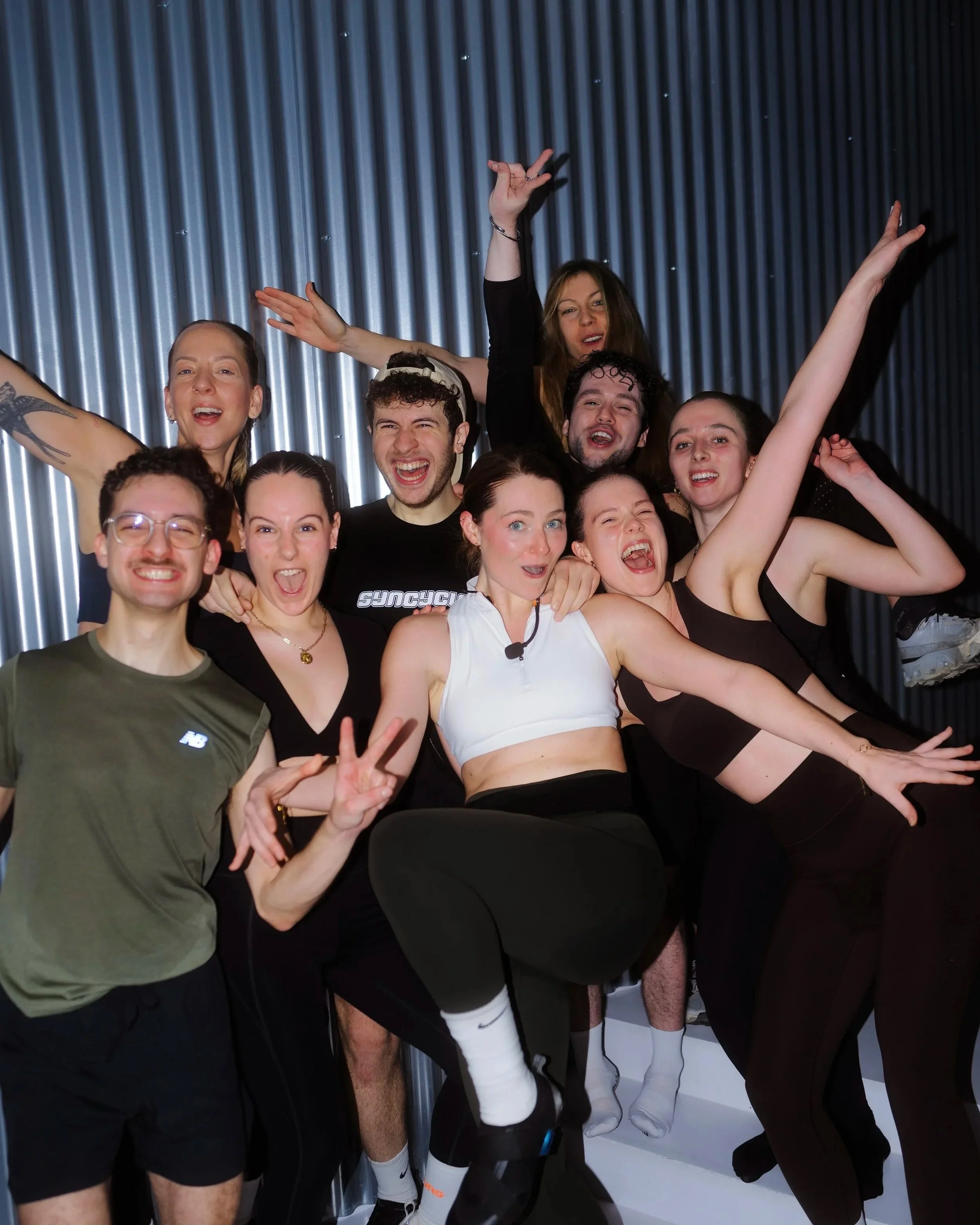 A group of people celebrating, smiling, and raising their hands in a gym or sports setting with a metallic corrugated wall behind them.