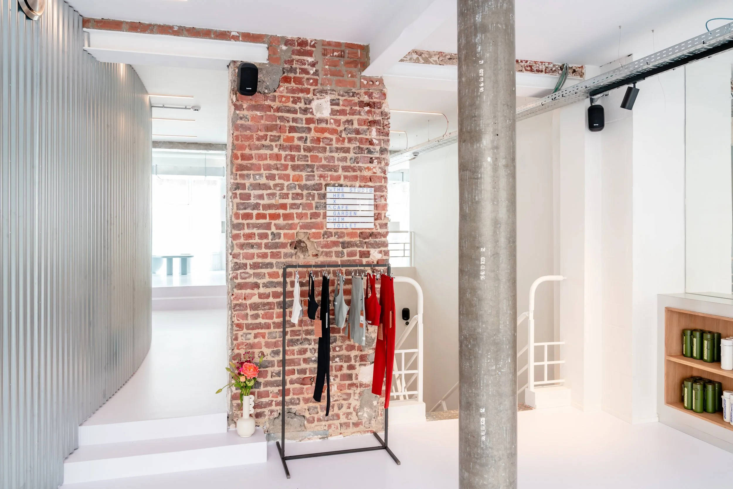 Interior view of a modern space with a brick wall, clothing rack with hanging clothes, a vase with pink flowers, and signage on the brick wall indicating different rooms or areas.