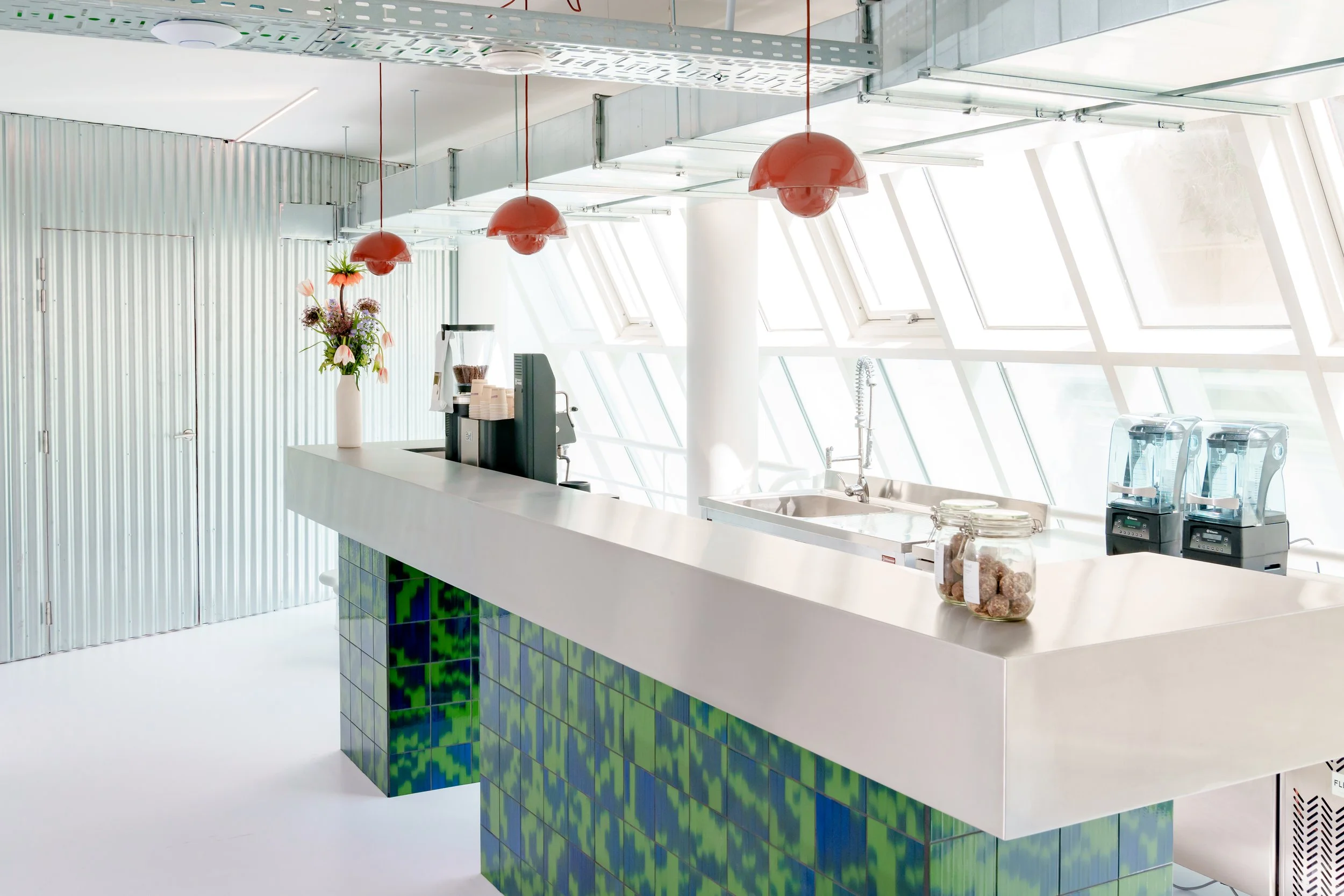 Modern coffee shop with a white counter, green tiled base, and large windows allowing natural light, red pendant lights, a vase with flowers, coffee machines, and jars with baked goods.