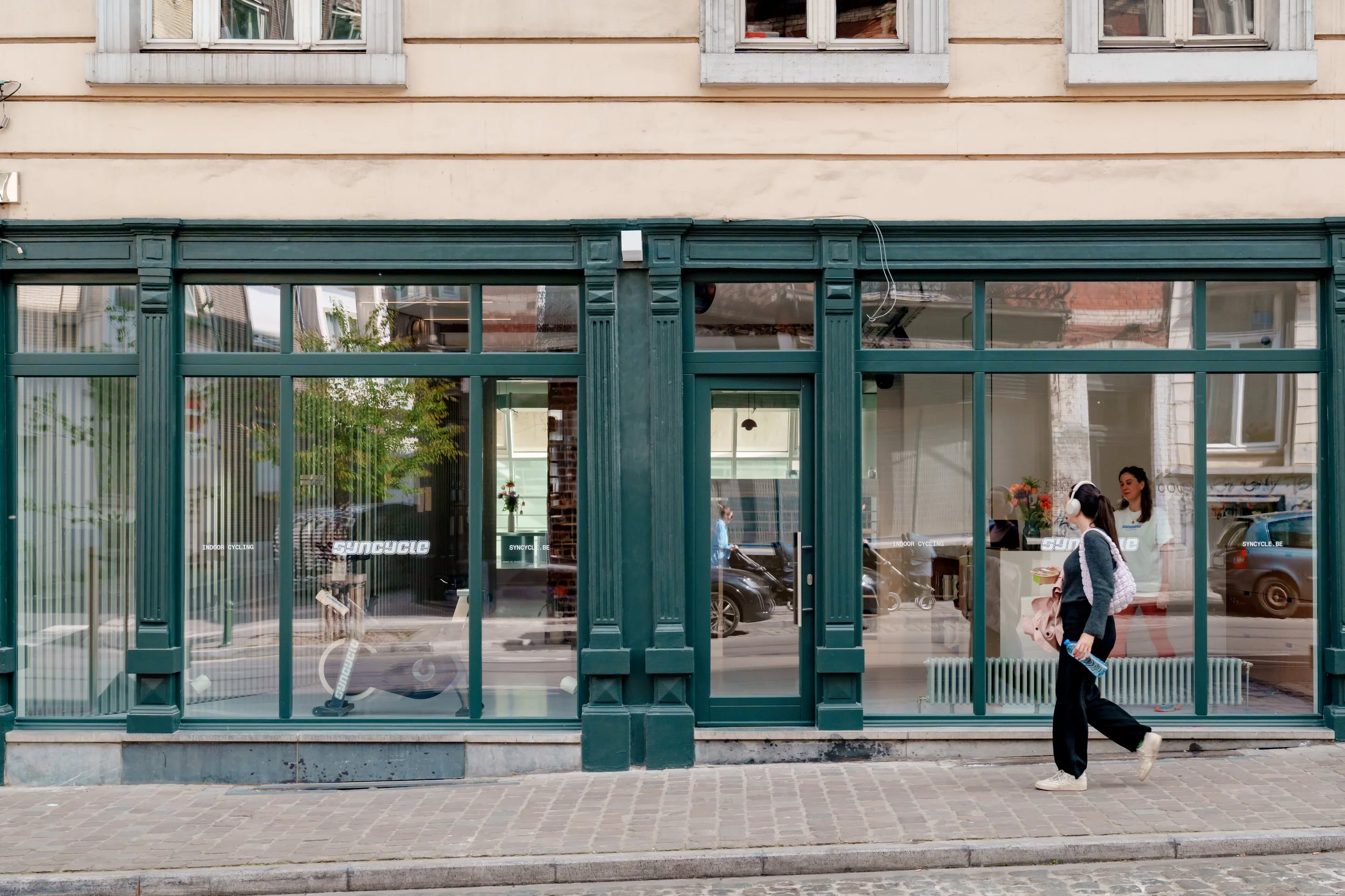 A woman in a hoodie and headphones walking past a gym with large glass windows in an urban setting.