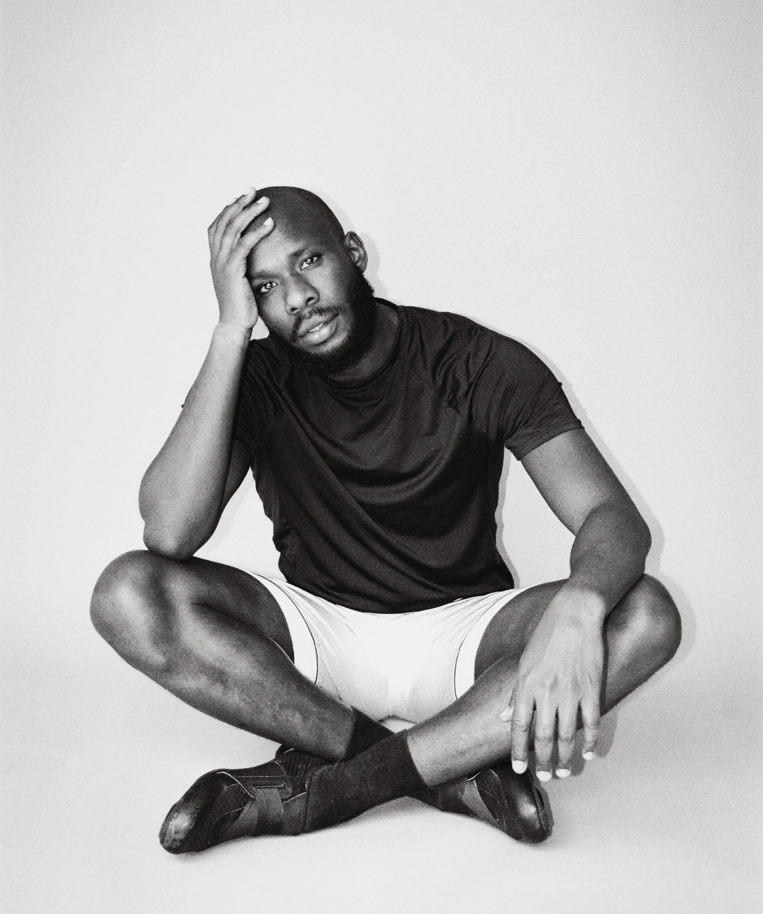 A black-and-white photo of a young man sitting on the floor against a plain background, wearing a dark t-shirt, white shorts, and black shoes, with one hand on his head and the other on his knee, looking directly at the camera.