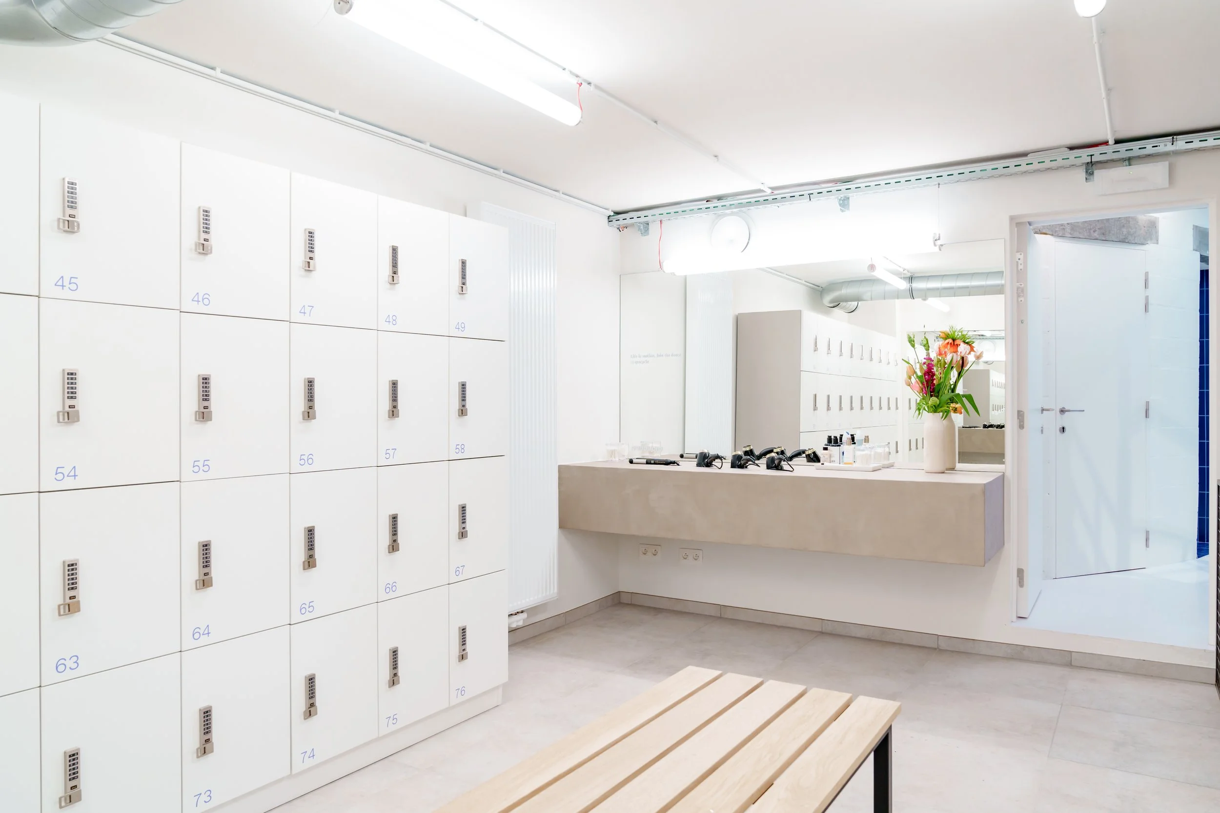 First aid station with white lockers numbered 45 to 76 on the left, a seating bench in the foreground, and a counter with black massage rollers and toiletries beside a large vase of flowers, located in a bright, modern room.