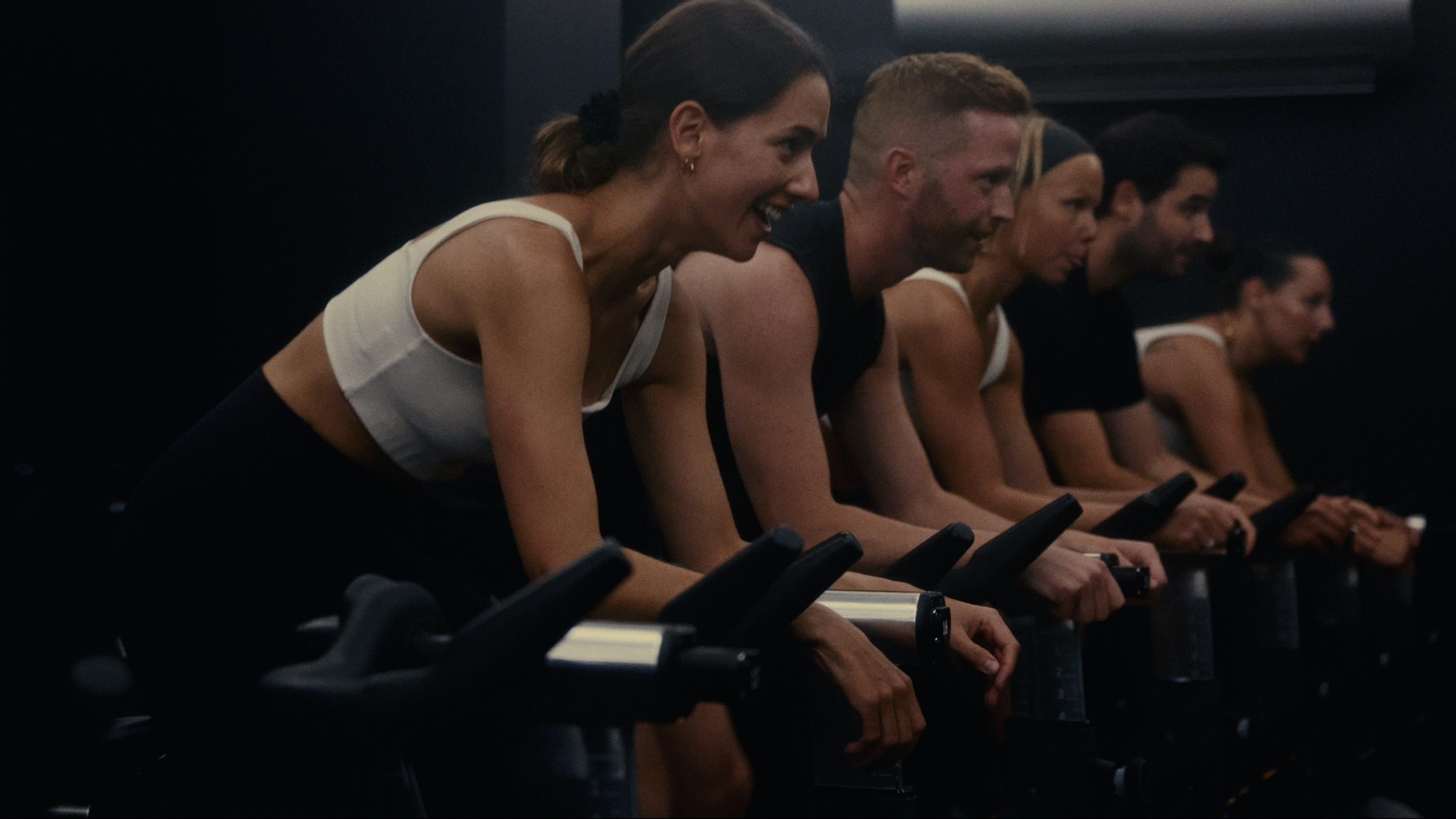 Five people in sportswear participating in a spin class, pedaling exercise bikes in a dark room.