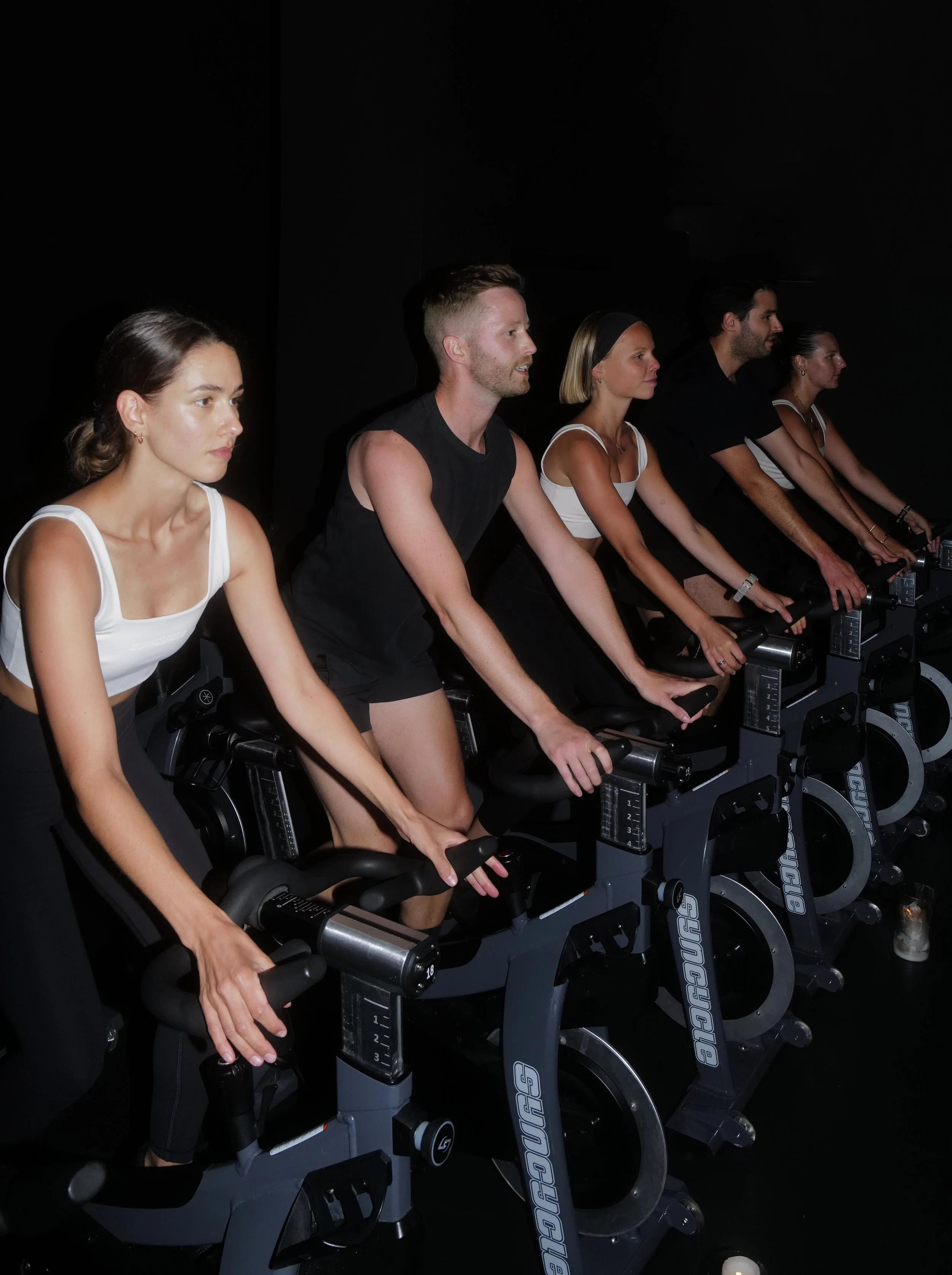Six people participating in a spin class, riding stationary bikes in a dark room.