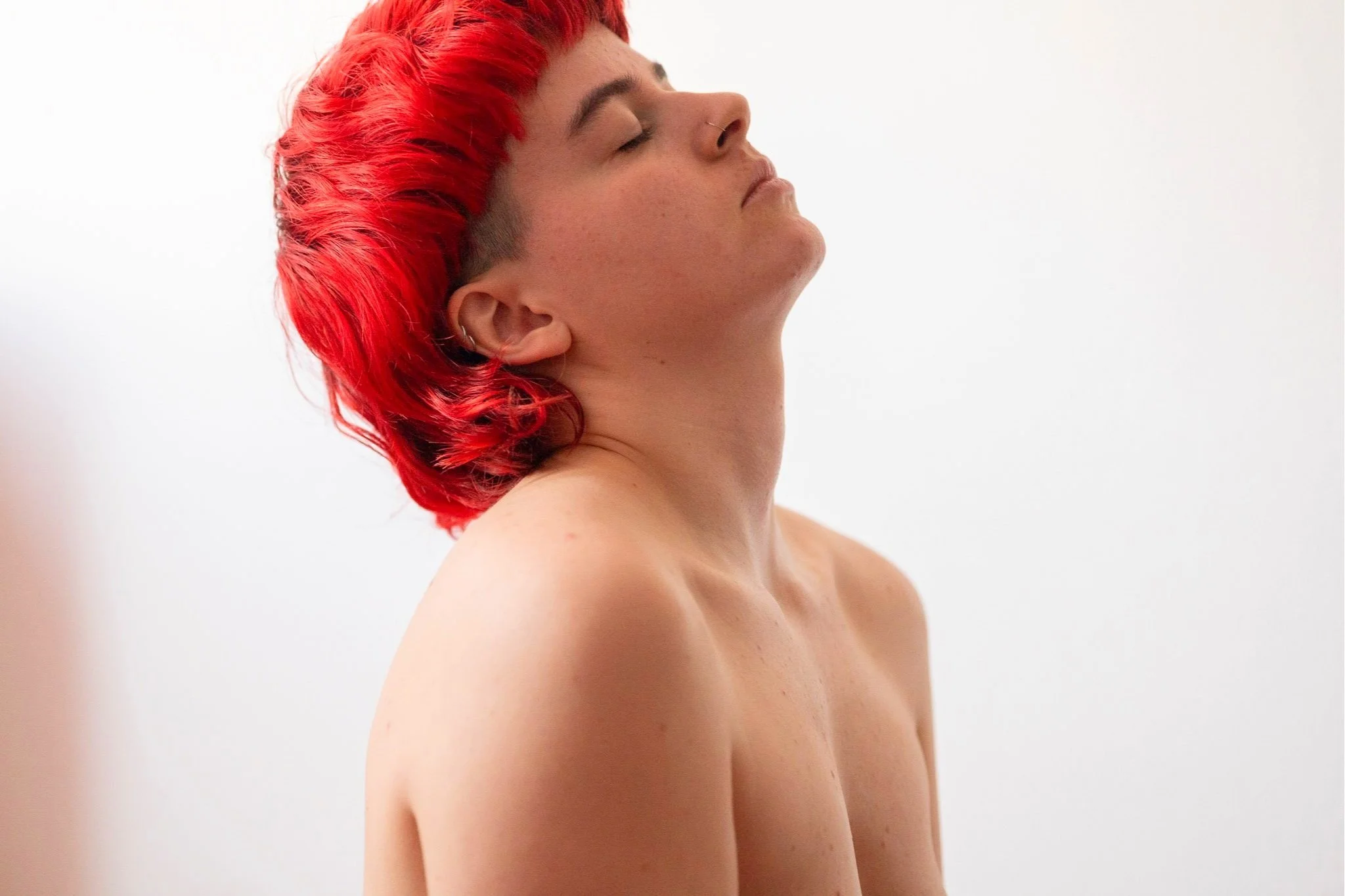 Nahaufnahme einer nackten Frau mit kurzen roten Haaren, die die Augen geschlossen hat und den Kopf leicht nach hinten neigt, vor hellem Hintergrund.