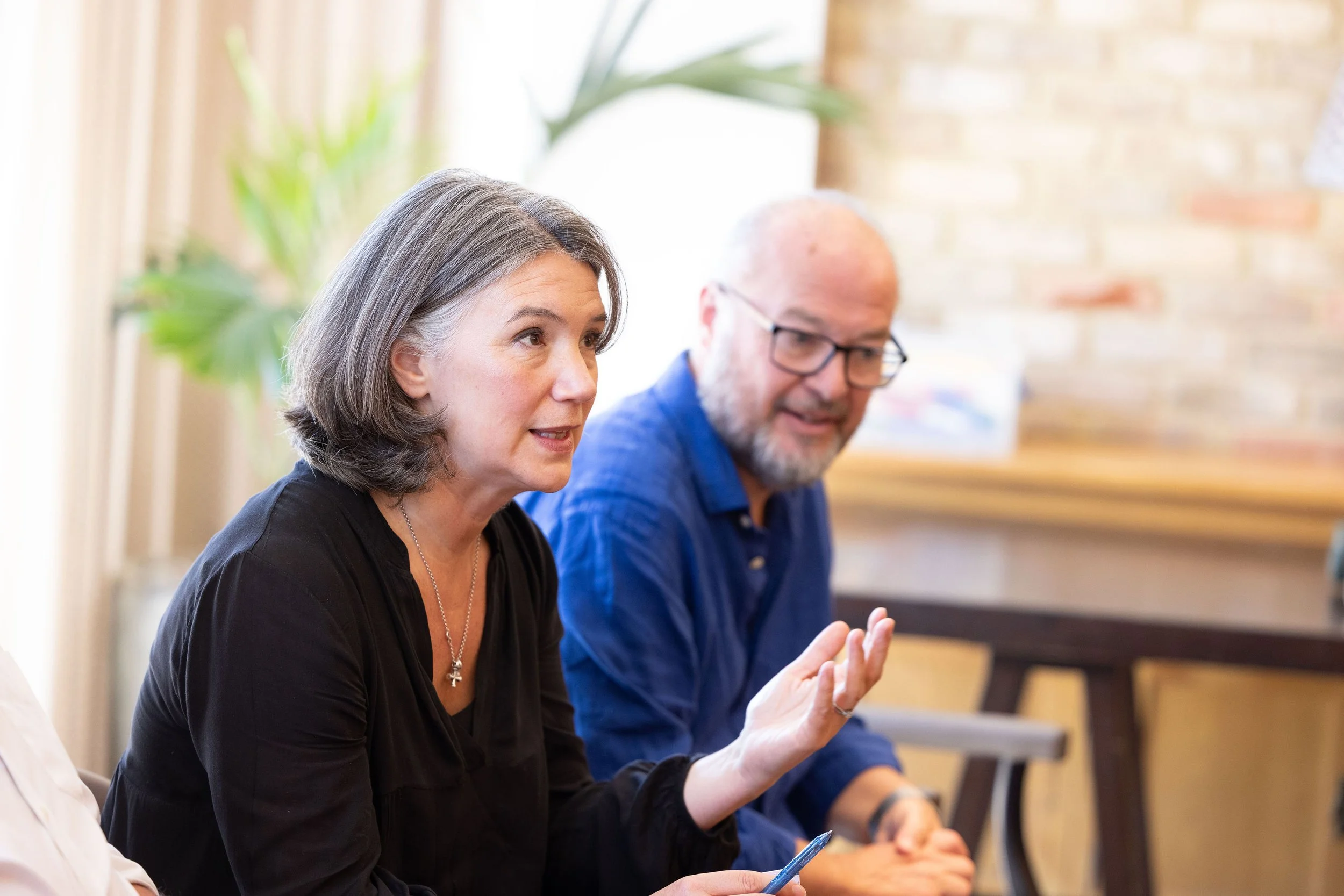 Rebecca Rumsey, executive and leadership coach, speaking and gesturing during a meeting, sitting beside a man with glasses listening. They are in a bright room with a brick wall and a plant in the background.