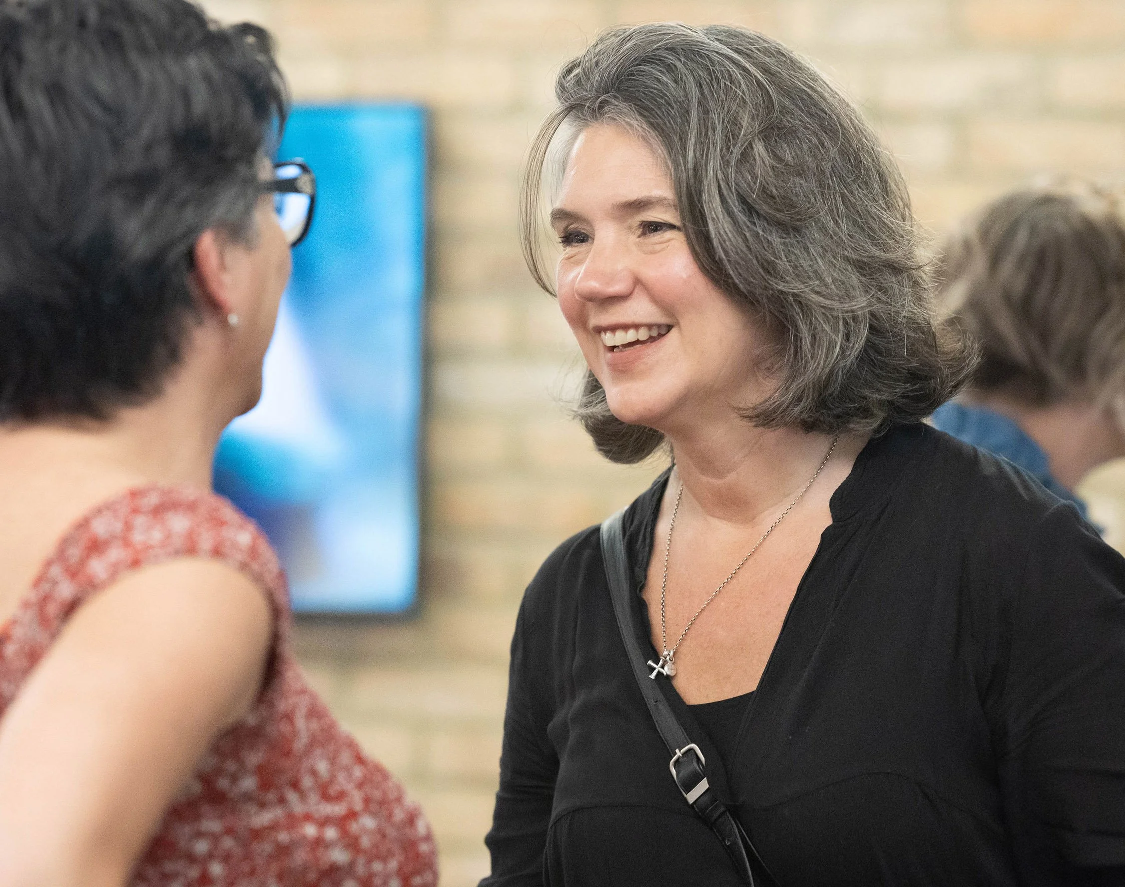 Rebecca Rumsey, executive and leadership coach, talking with a client at a conference, with a blurred background of another person and a brick wall.
