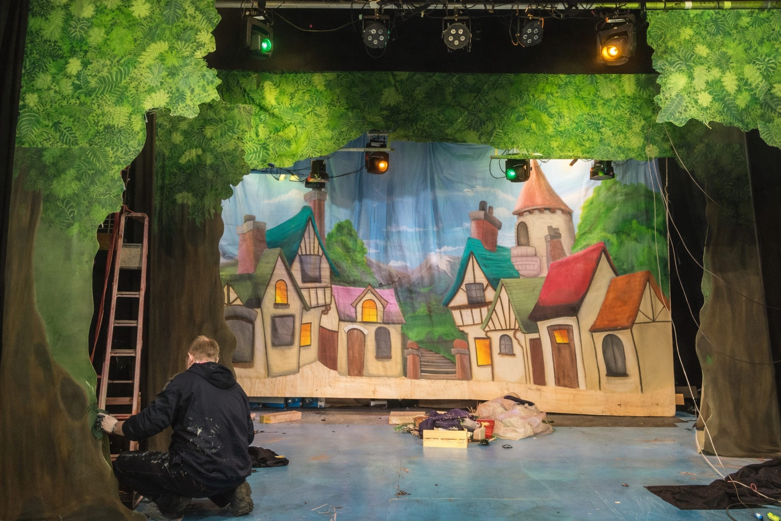 Stage set being painted to resemble a fairy tale village with cottages, a castle, and trees, with a person working on the scenery. Pantomime set design.