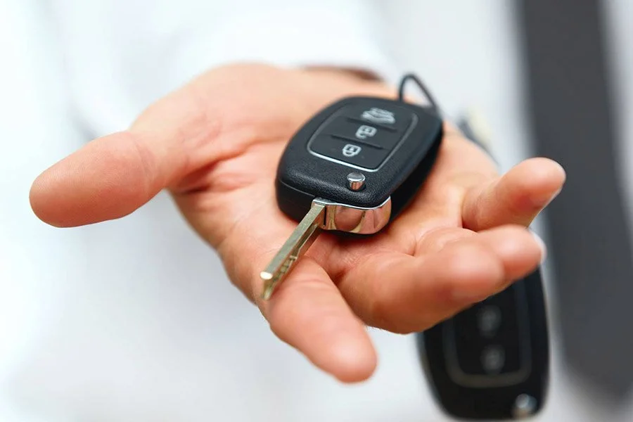 Close-up of a person's hand holding a car key fob with buttons for lock, unlock, and trunk, with a car door in the background.
