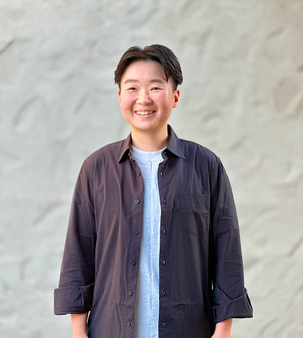 Young person smiling in casual attire, standing outdoors against a light-colored wall.