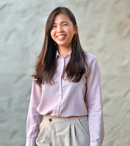 A young woman with long dark hair smiling, wearing a light pink button-up shirt and beige pants, standing outdoors with a blurred stone wall background.