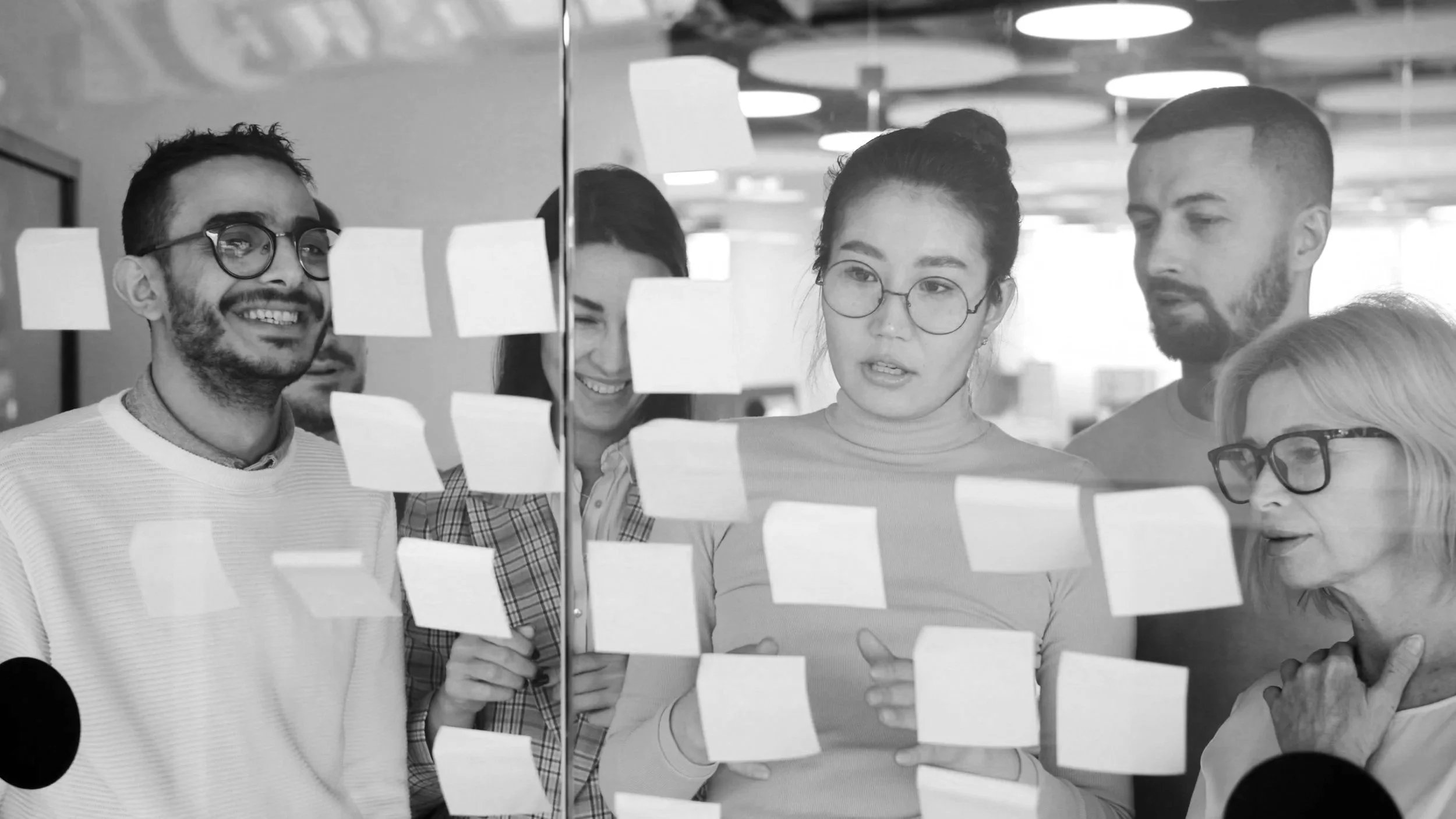 Five diverse people looking at sticky notes on a glass wall during a team meeting or brainstorming session.