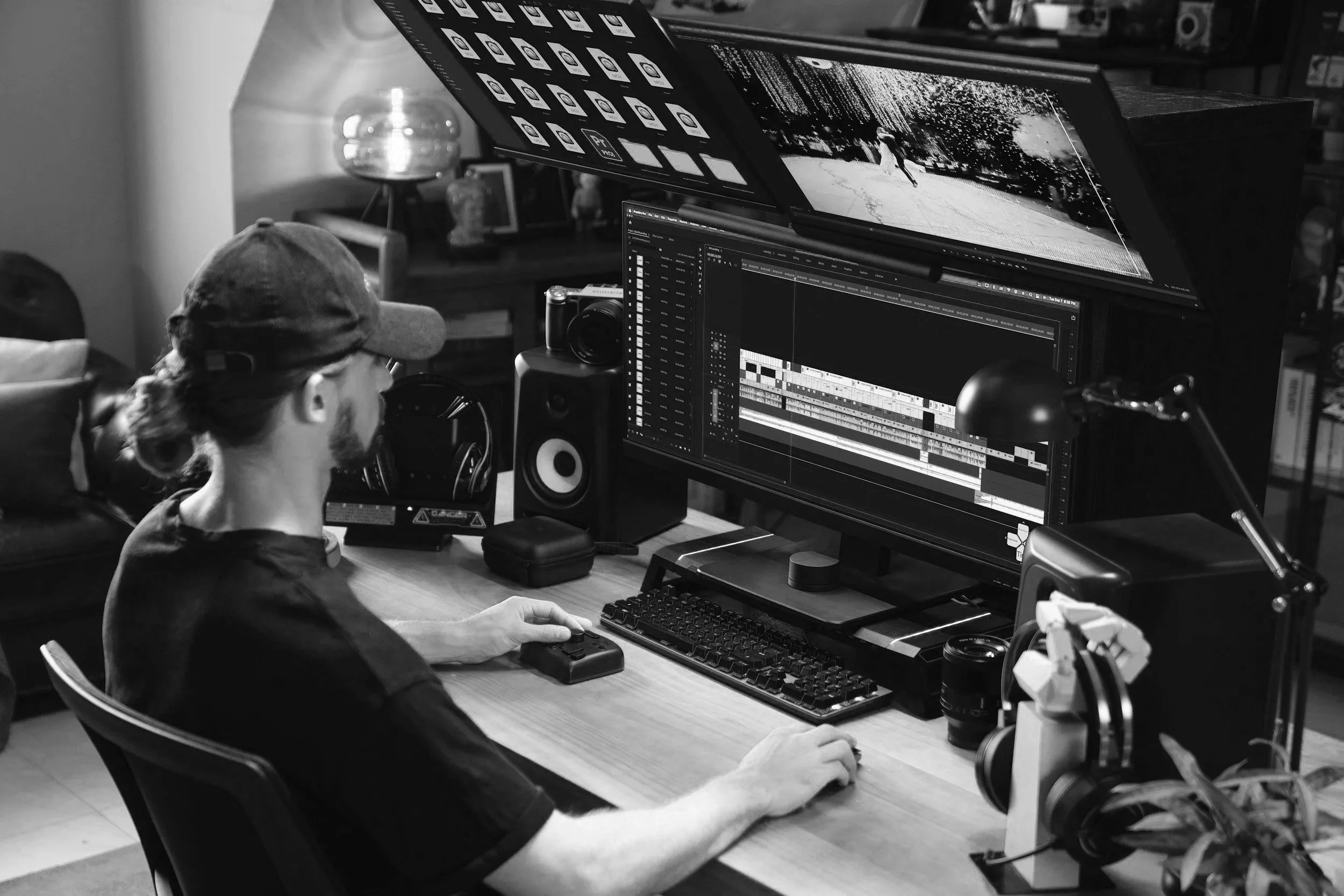 A person working on video editing at a desk with multiple monitors displaying editing software, surrounded by audio equipment and personal items.