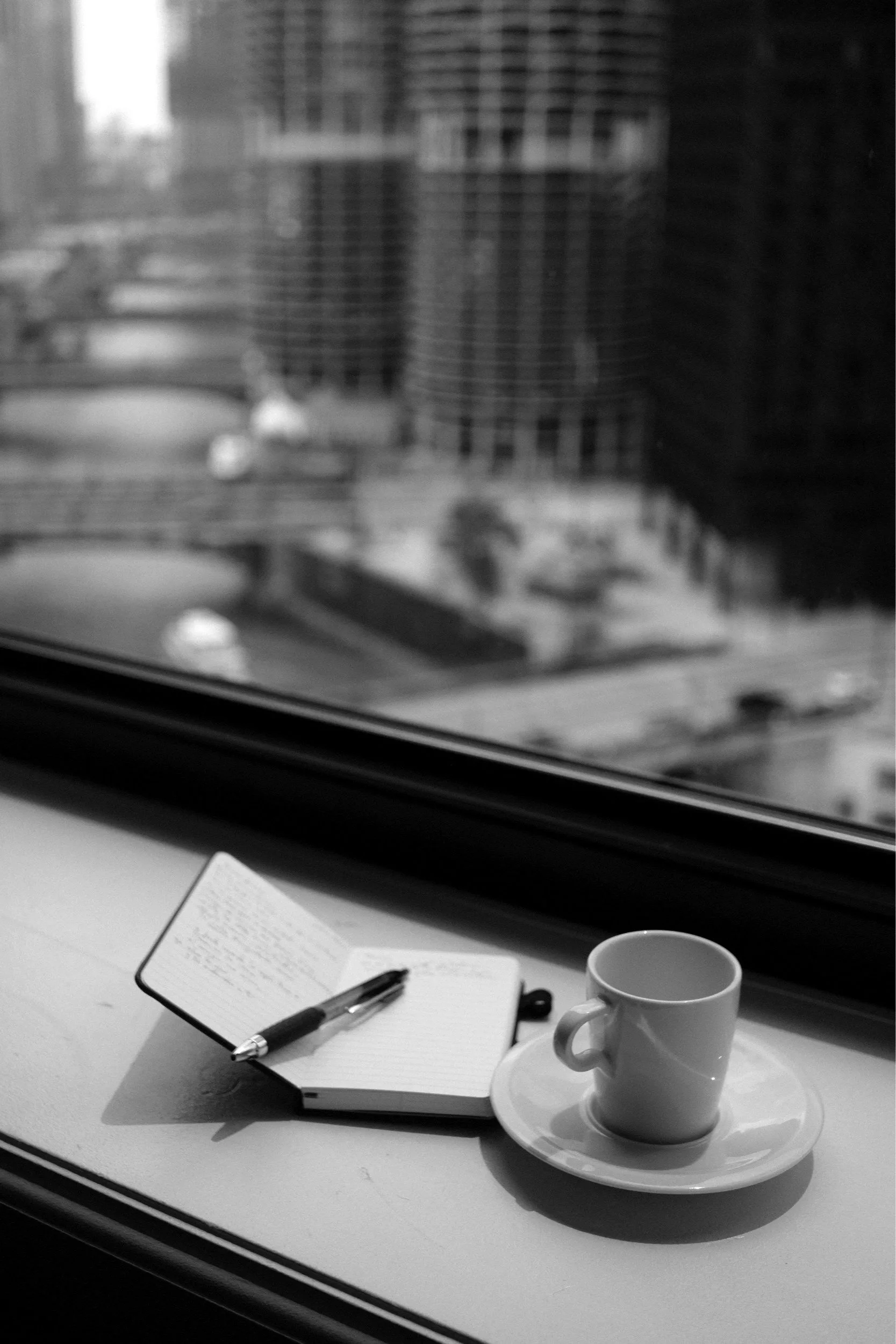 A black and white photograph of a window ledge with an open notebook, a pen, and a coffee cup with a saucer, with city buildings and blurred urban scenery outside the window.