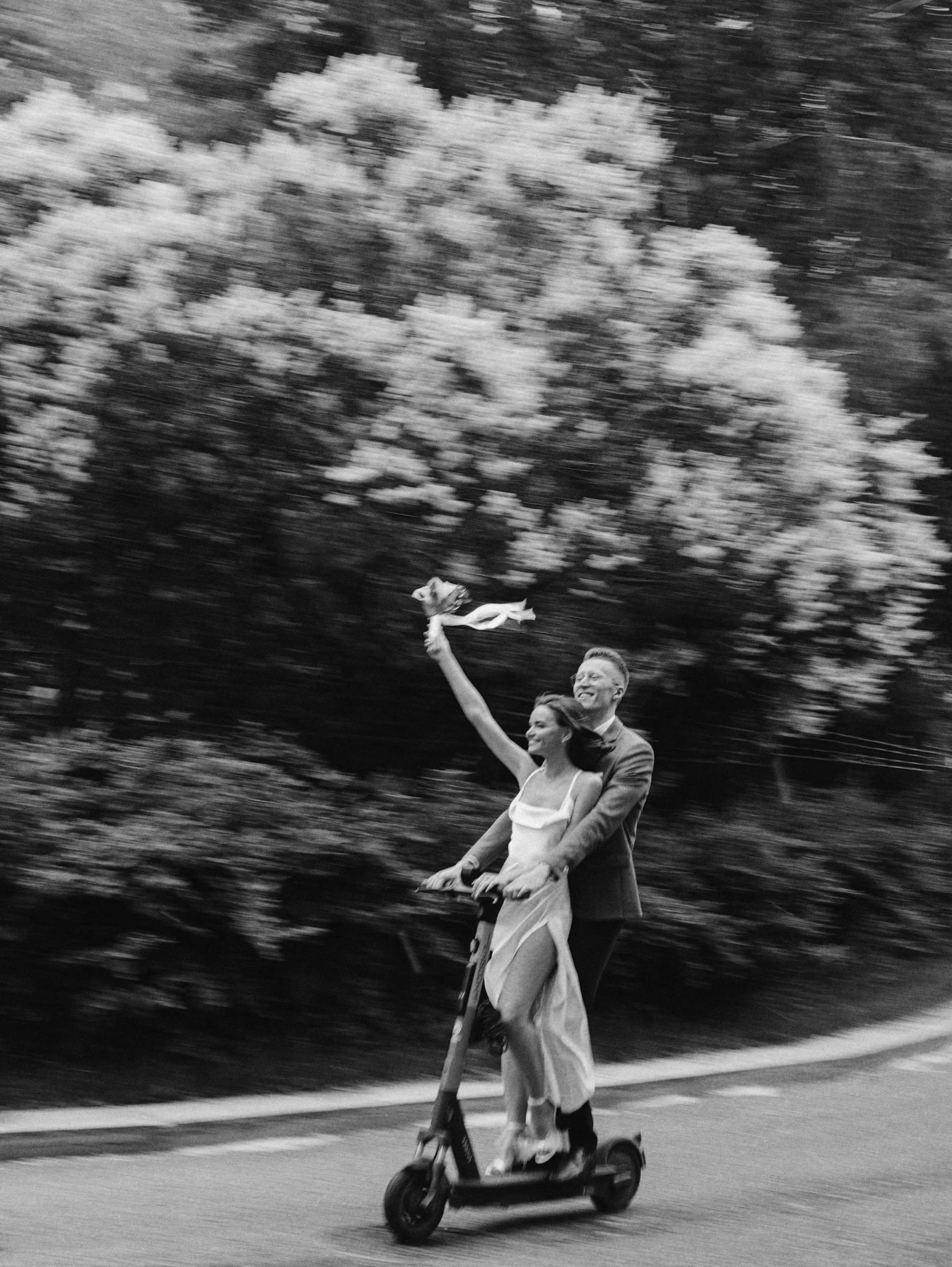 A couple riding an electric scooter on a road, with the woman sitting in front holding a bouquet, smiling, and waving, while the man behind her rides and holds her around the waist. The background is blurred with trees.