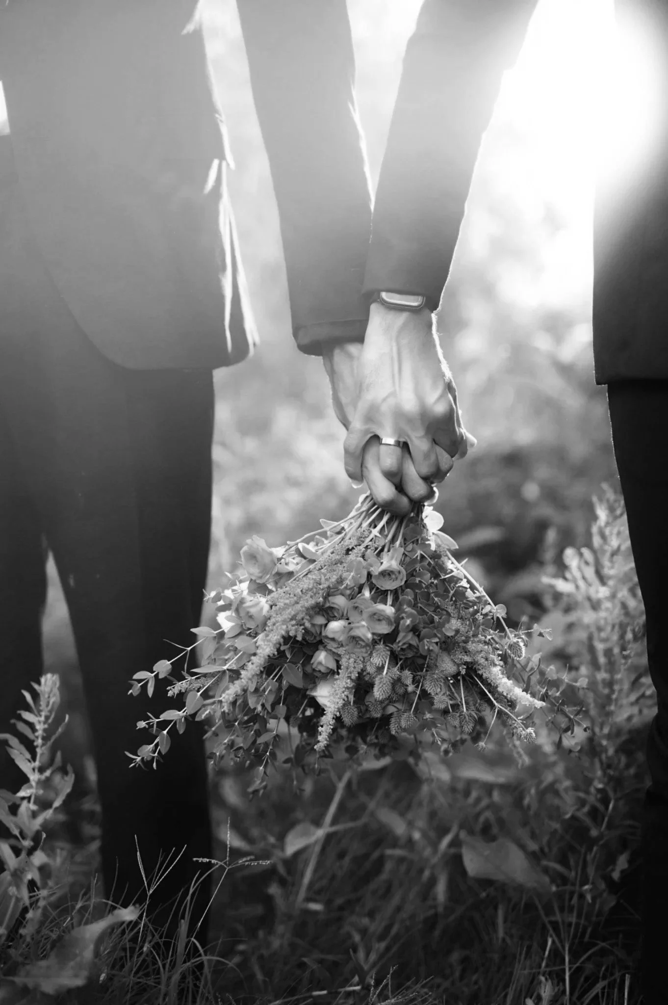 Two people holding hands, one of them holding a bouquet of flowers, outdoors in a natural setting, in black and white.