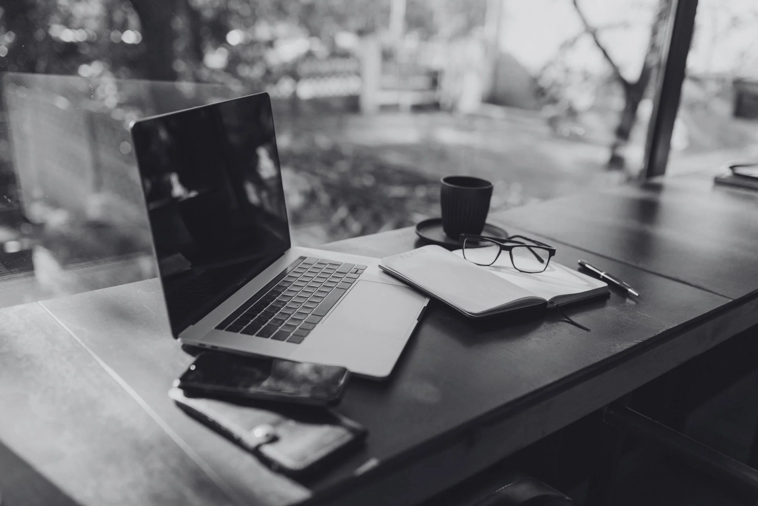 A desk with an open laptop, eyeglasses, notebook, pen, smartphone, and a cup on a saucer, situated in front of a window with a view of trees and a yard in black and white.