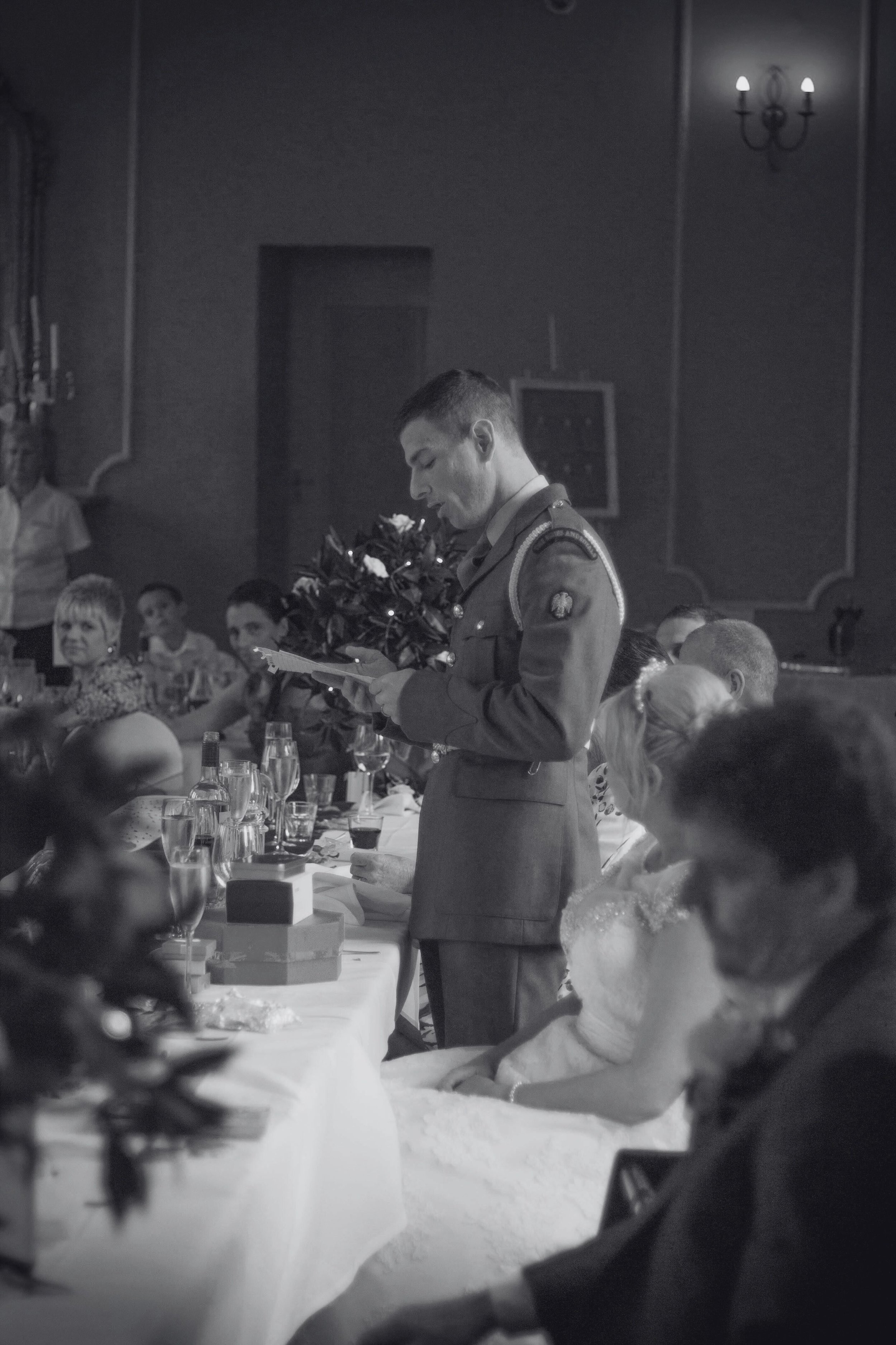 A man in a military uniform reading a card or speech at a formal event, surrounded by seated guests dressed in formal attire, at a decorated banquet table with wine glasses and flowers.
