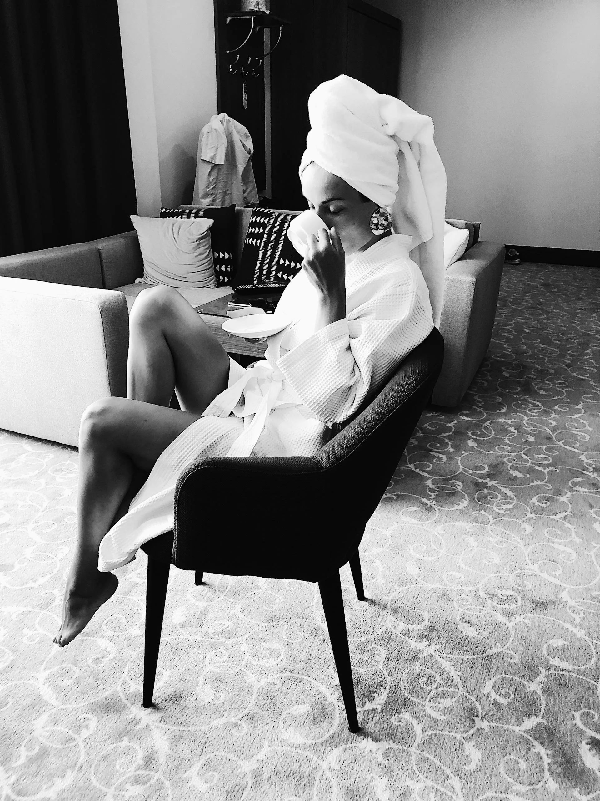 A woman with a towel wrapped around her head, wearing a bathrobe, sitting on a black chair and drinking tea or coffee in a hotel room.