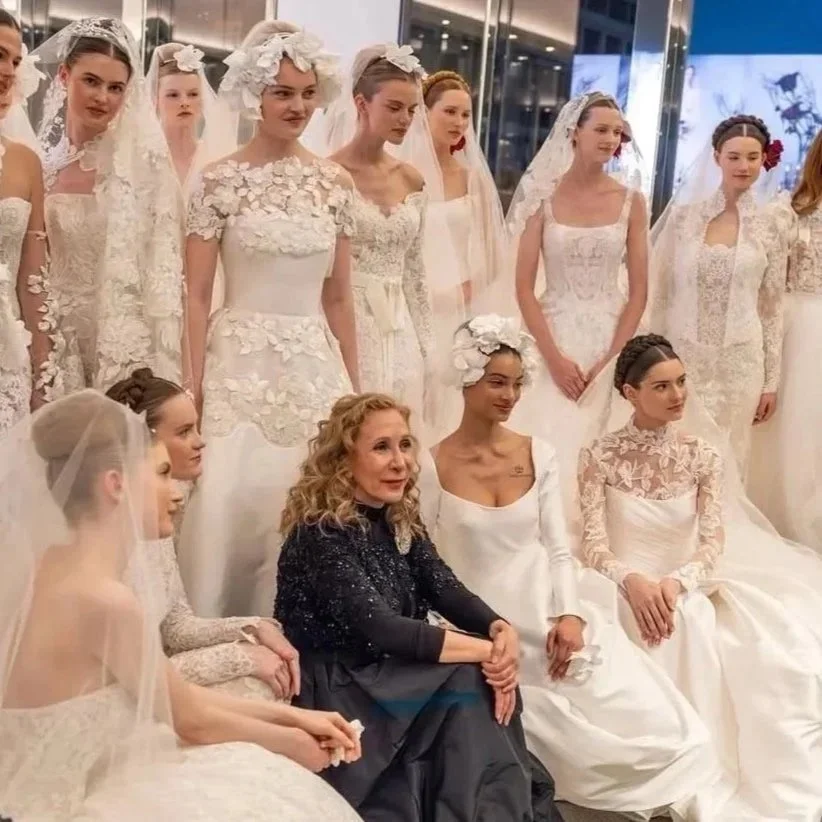 A group of women in white wedding dresses, with one woman in a dark dress, posing for a photo in what appears to be a bridal boutique or showroom.