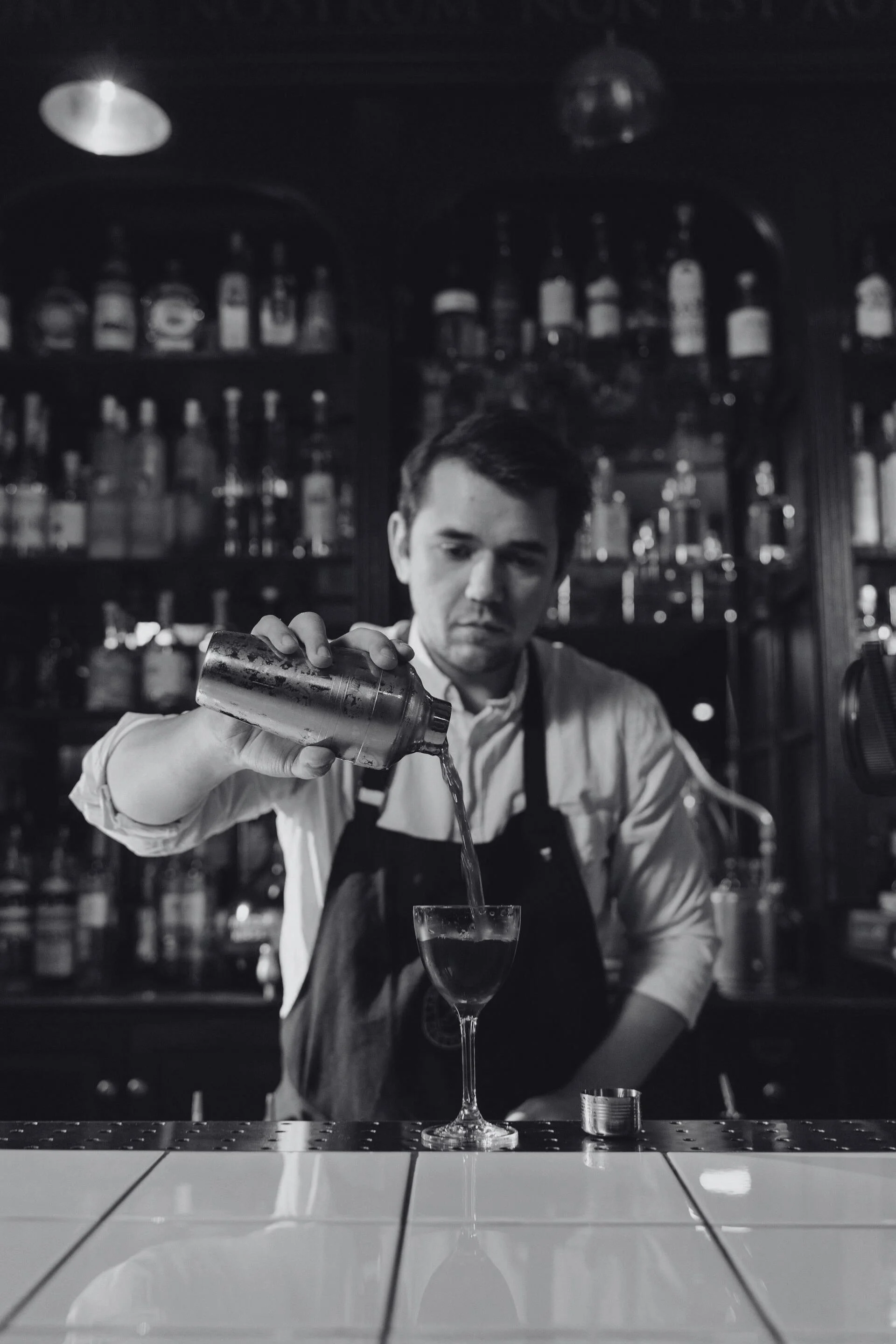 A bartender pouring a drink into a coupe glass behind a bar with bottles on shelves in the background.