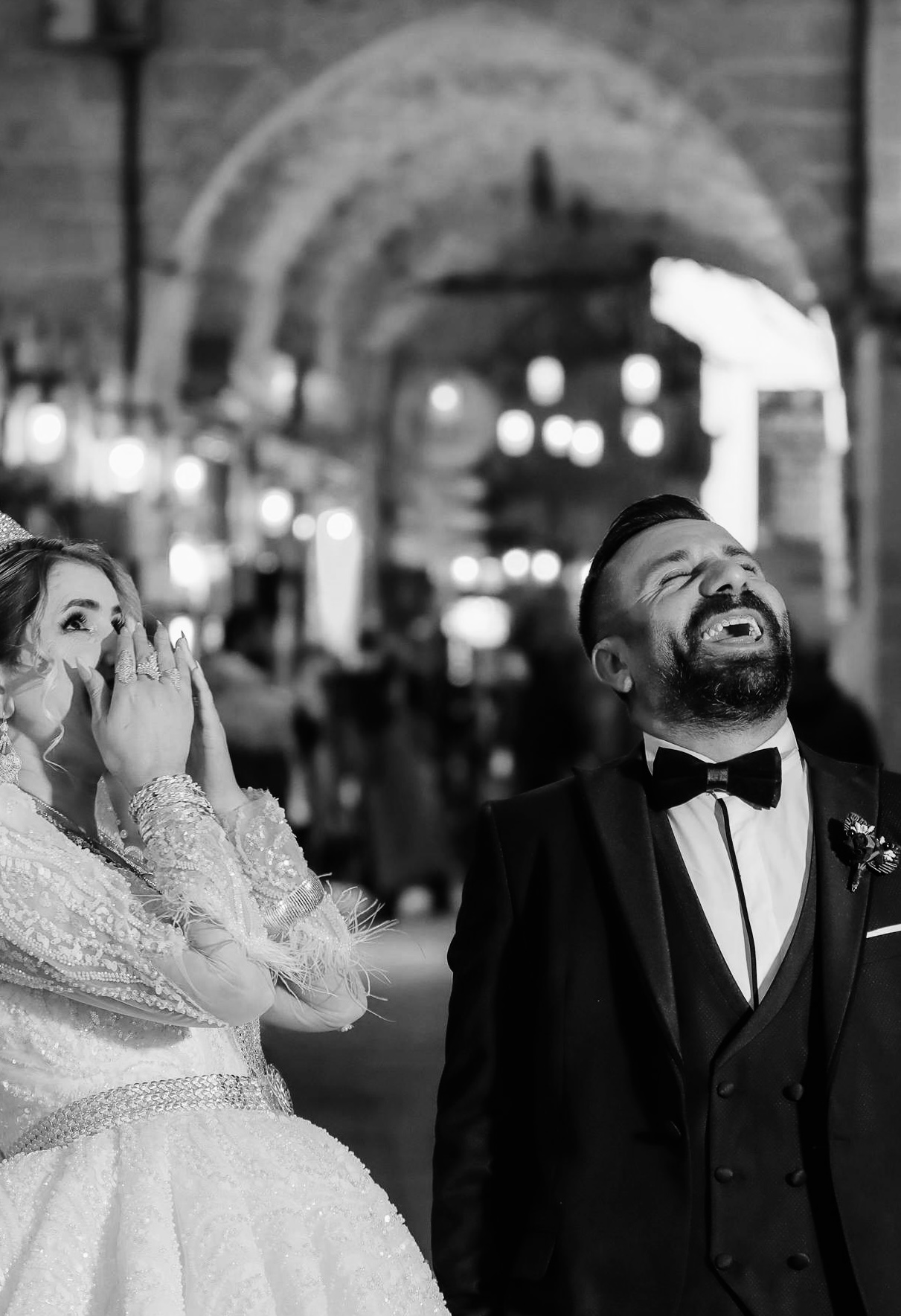 A black and white photo of a bride and groom on their wedding day, with the bride covering her mouth in surprise or joy, and the groom smiling or laughing with closed eyes, in an elegantly decorated venue with hanging lights.