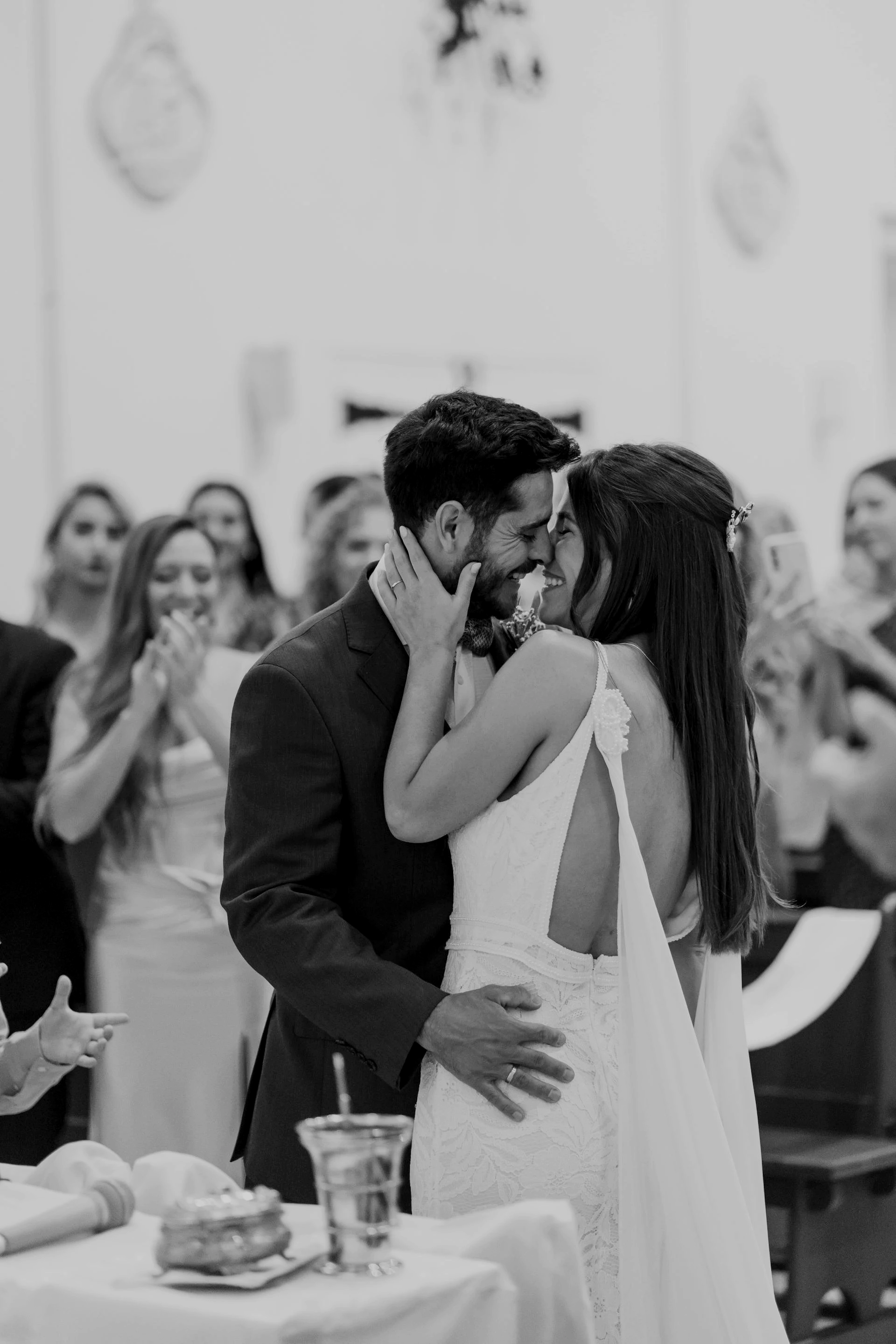 A couple is dancing closely at their wedding, smiling with their noses touching. They are surrounded by friends and family who are clapping and celebrating.
