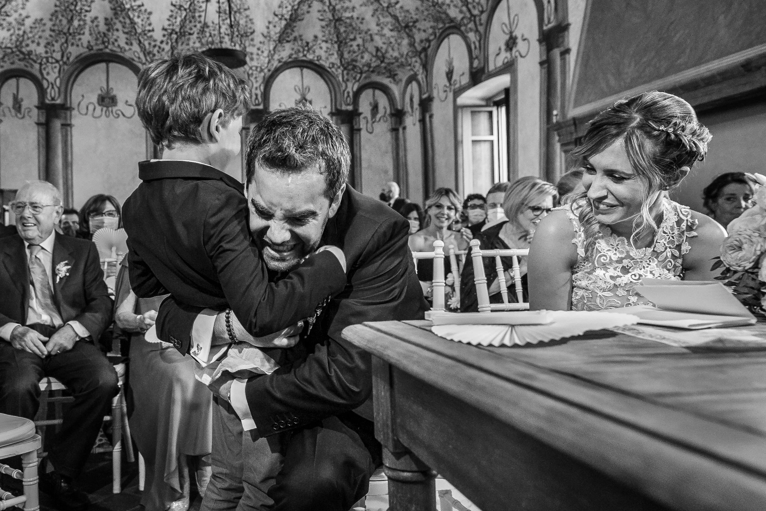 A man and a young boy are hugging during a wedding ceremony, with the man smiling through the embrace. A woman in a lace dress is watching and smiling, sitting at a table with documents and flowers. Guests are seated in a decorated hall with arched w