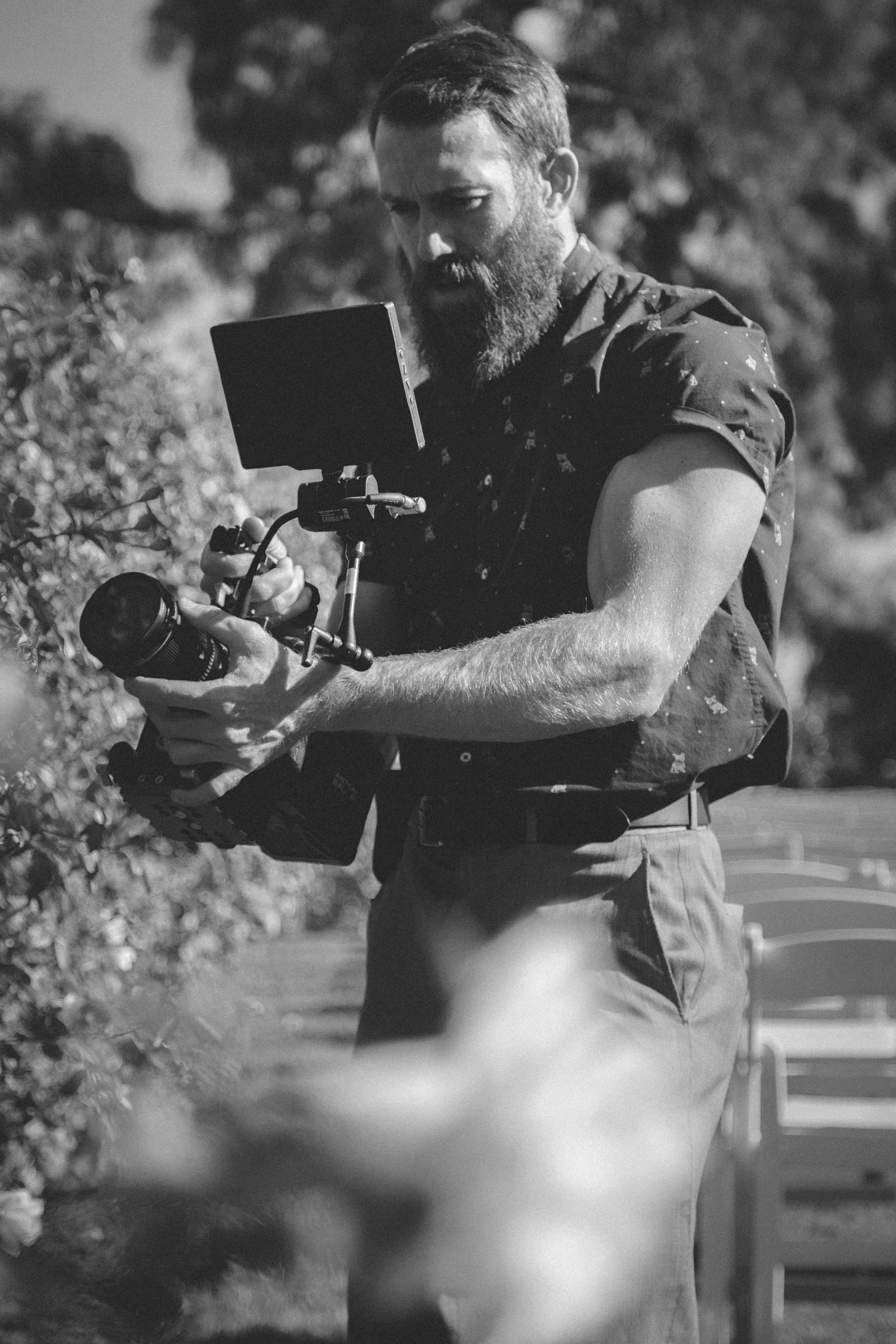 A man with a beard and short hair operating a camera mounted on a stabilizer outdoors in black and white.