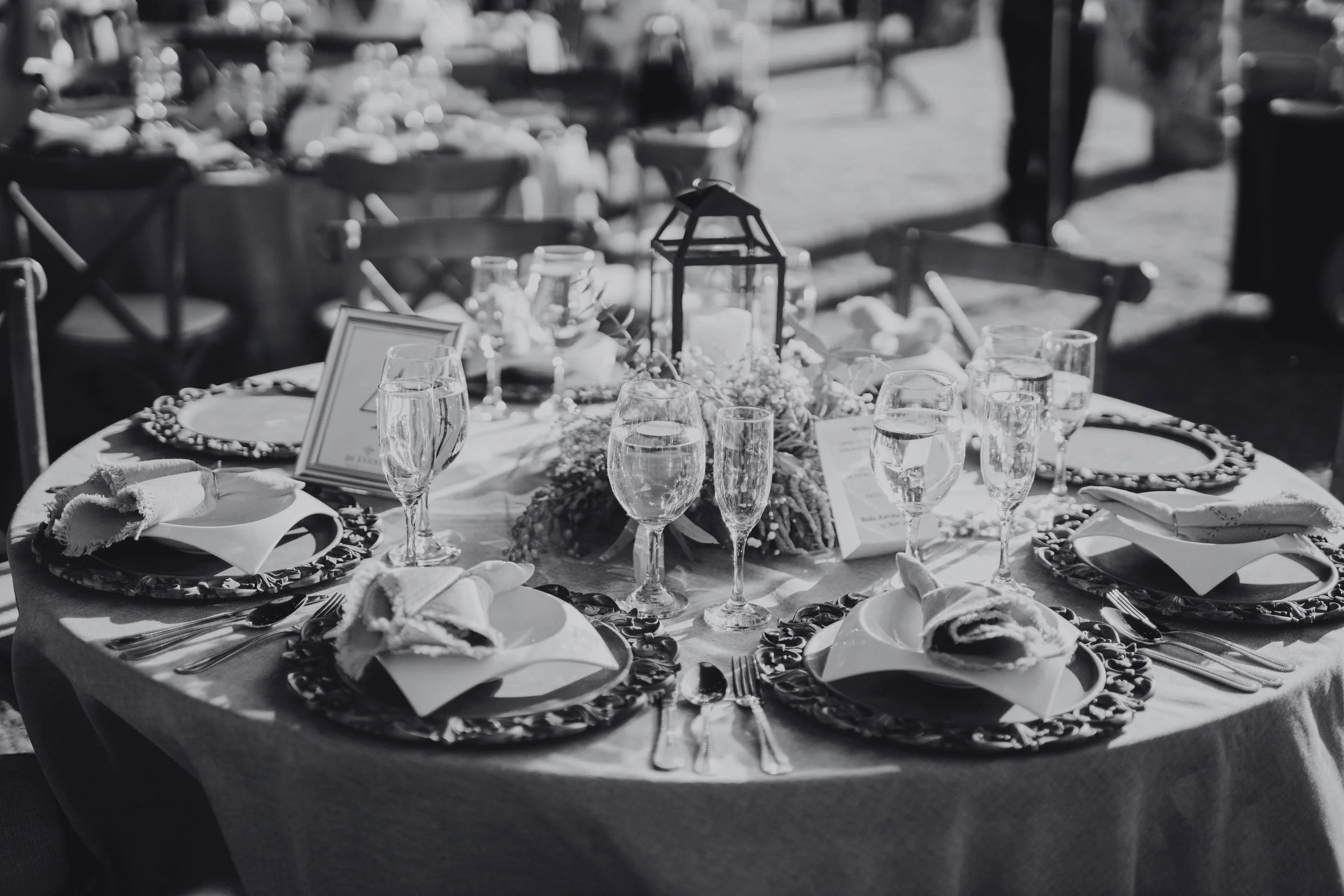 A round wedding reception table set with decorative plates, cloth napkins, wine glasses, and a lantern centerpiece, outdoors with other tables in the background.