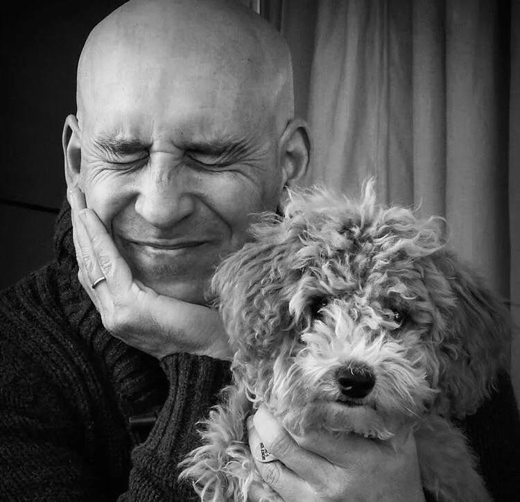 A man smiling with his eyes closed, holding a fluffy poodle puppy near his face. The man has a bald head and is wearing a dark sweater, with a ring visible on his finger. The puppy has curly fur and is looking ahead, with a curtain in the background.