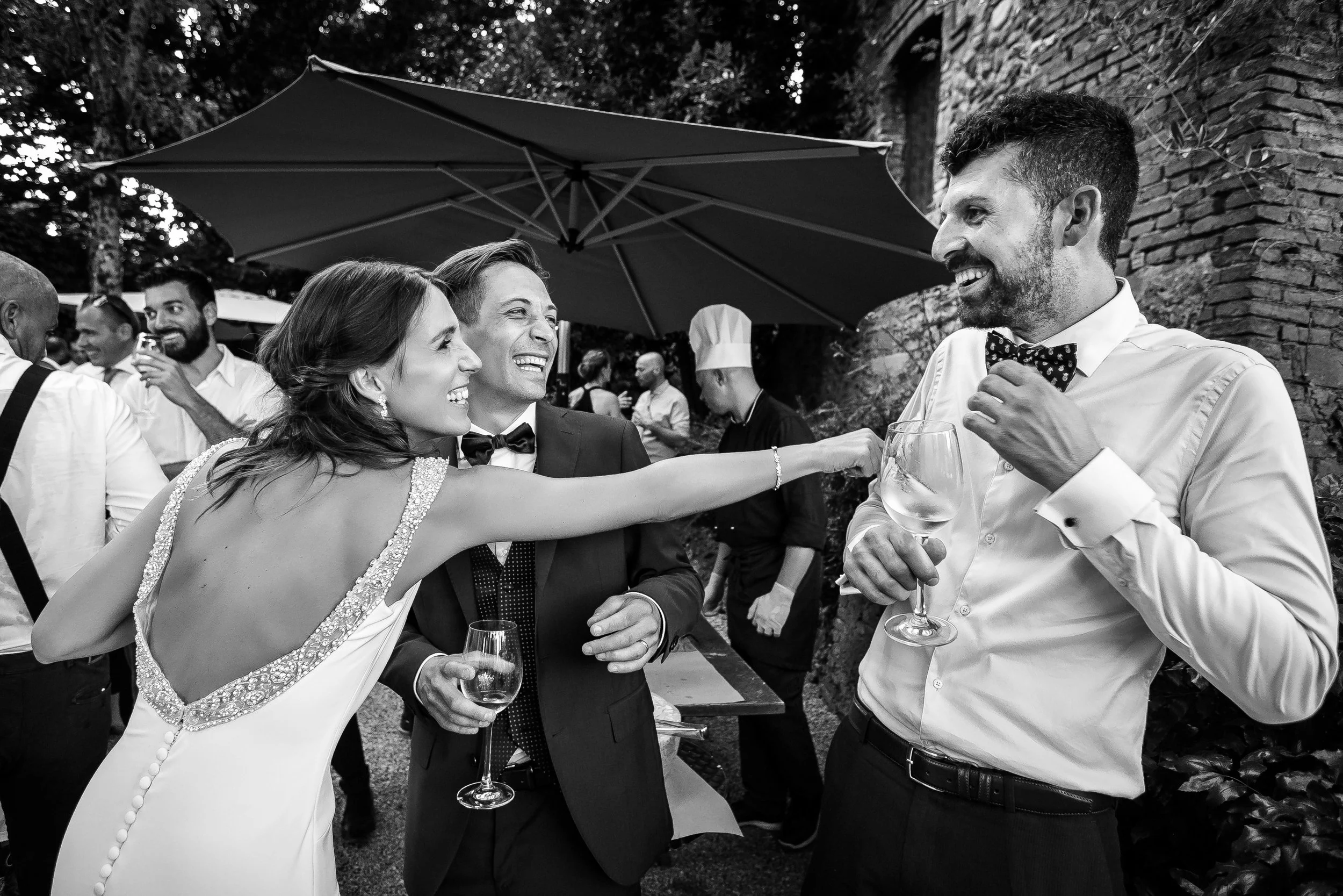 People at a social gathering, one woman in a dress playfully pointing at a man in a dress shirt and bow tie, with others smiling in the background.
