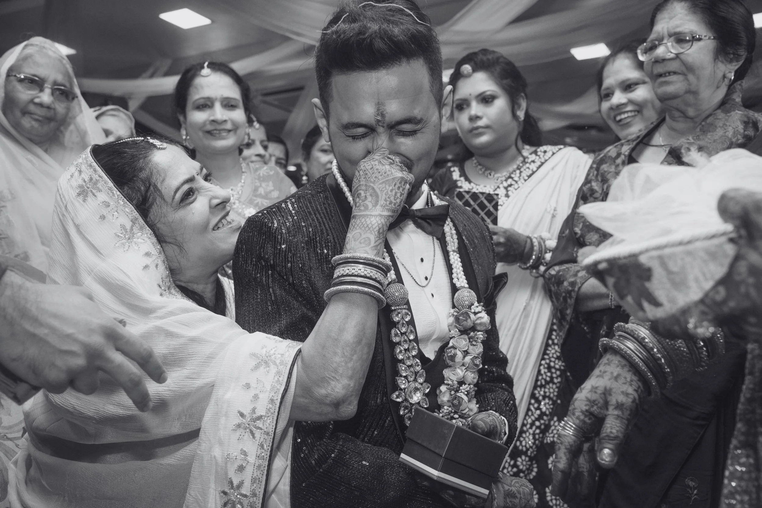 A bride and groom during their wedding, with the bride applying vermilion to the groom's forehead. The groom is wearing traditional wedding attire with garlands and jewelry, and the bride is in a saree with a veil. Family members surround them, smili
