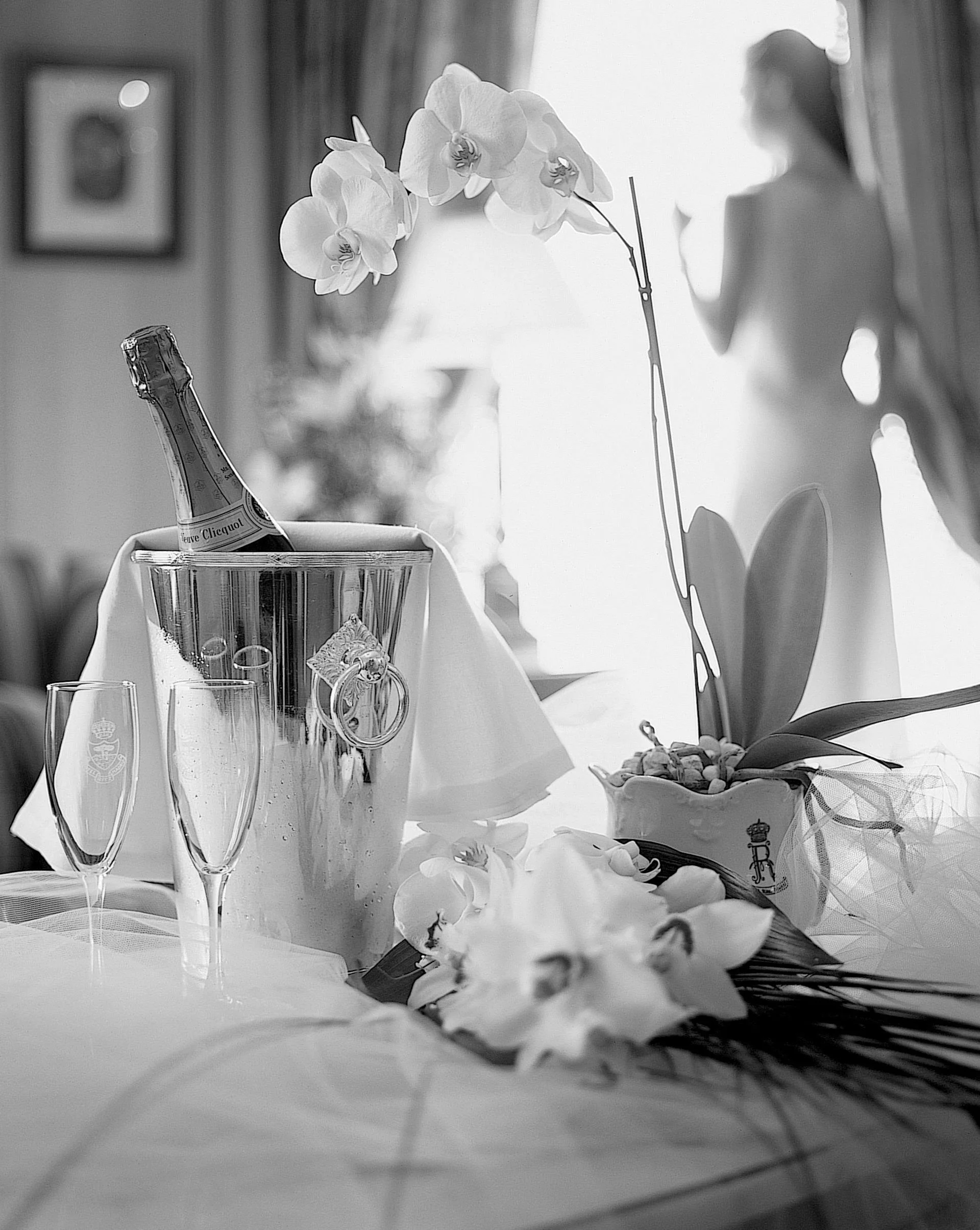 A black and white photo of a table set with flowers, champagne bottle in an ice bucket, empty champagne flutes, and a blurred angel figurine in the background.