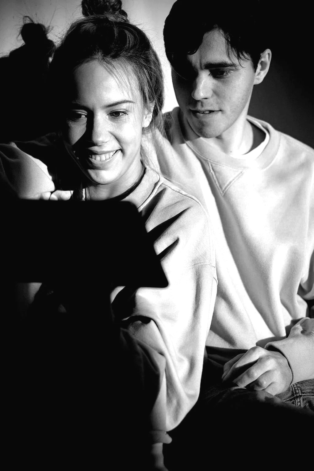 A black and white photo of a smiling young woman and a young man looking at a smartphone. The woman has her hair tied up in a bun, wearing a sweatshirt, and the man has dark hair, also in a sweatshirt. They are sitting close together, appearing engaged with what they are viewing.