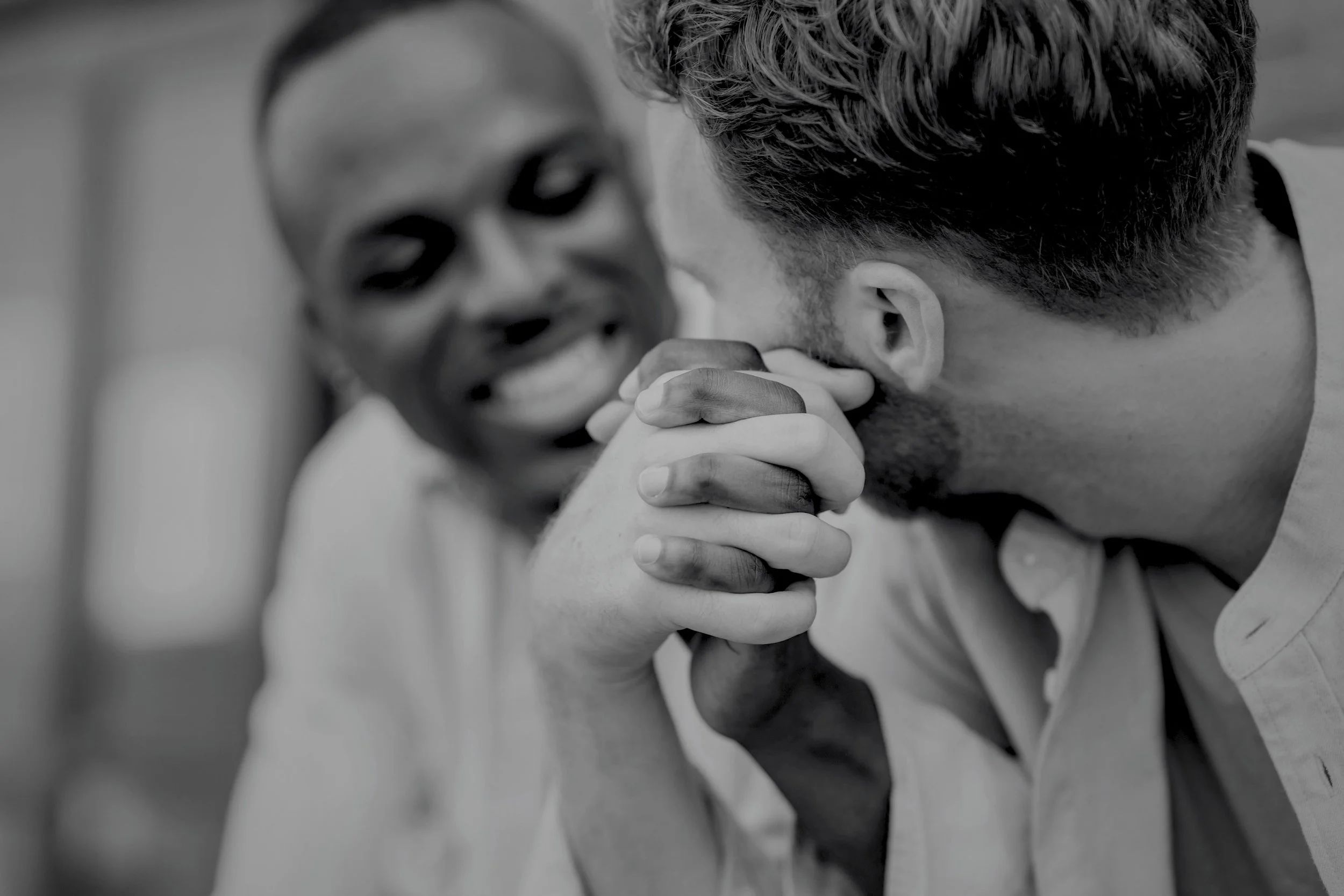 Two men are engaged in a handshake with their hands clasped, close to their faces, smiling and showing emotional connection.