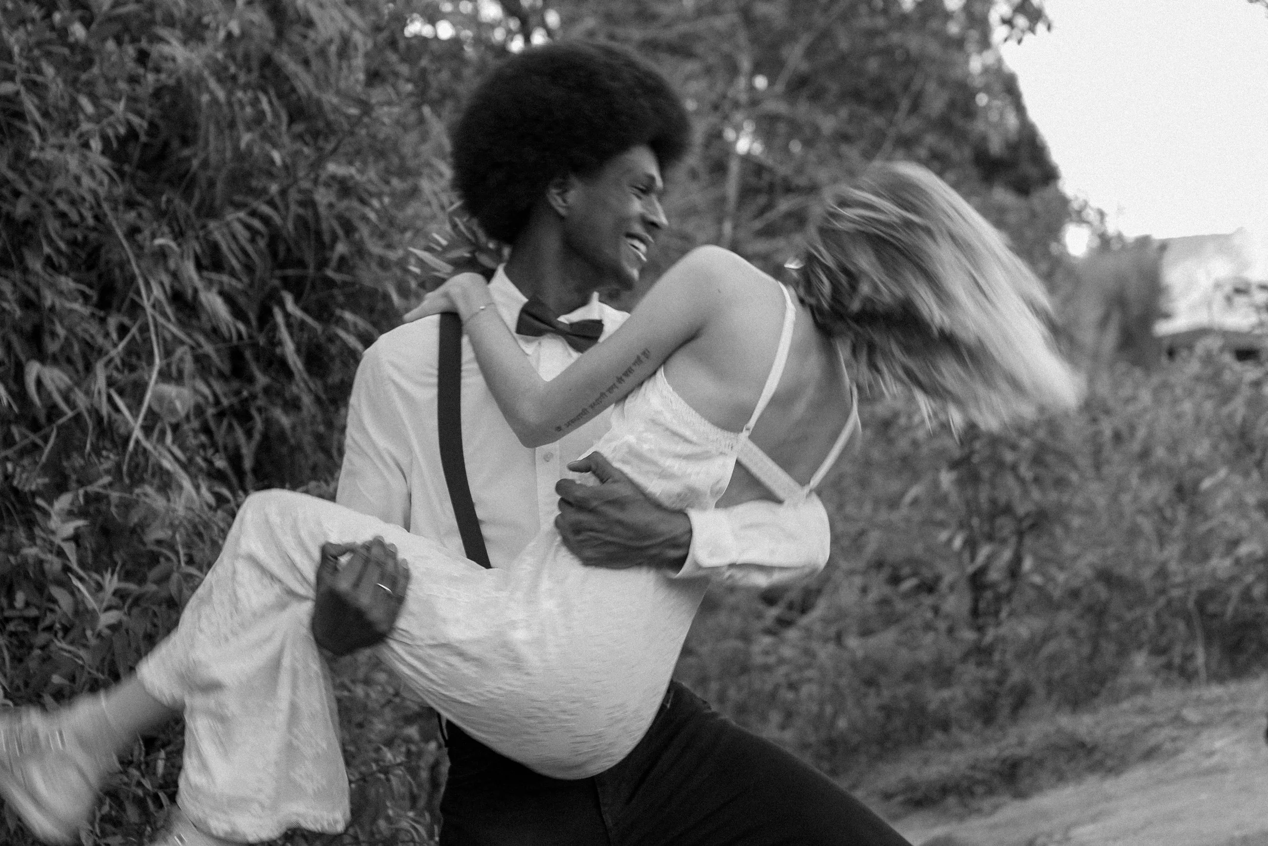 A man with an afro hairstyle and a woman with long, straight hair are outdoors. The man is smiling and holding the woman, who is wearing a dress, in a playful pose.