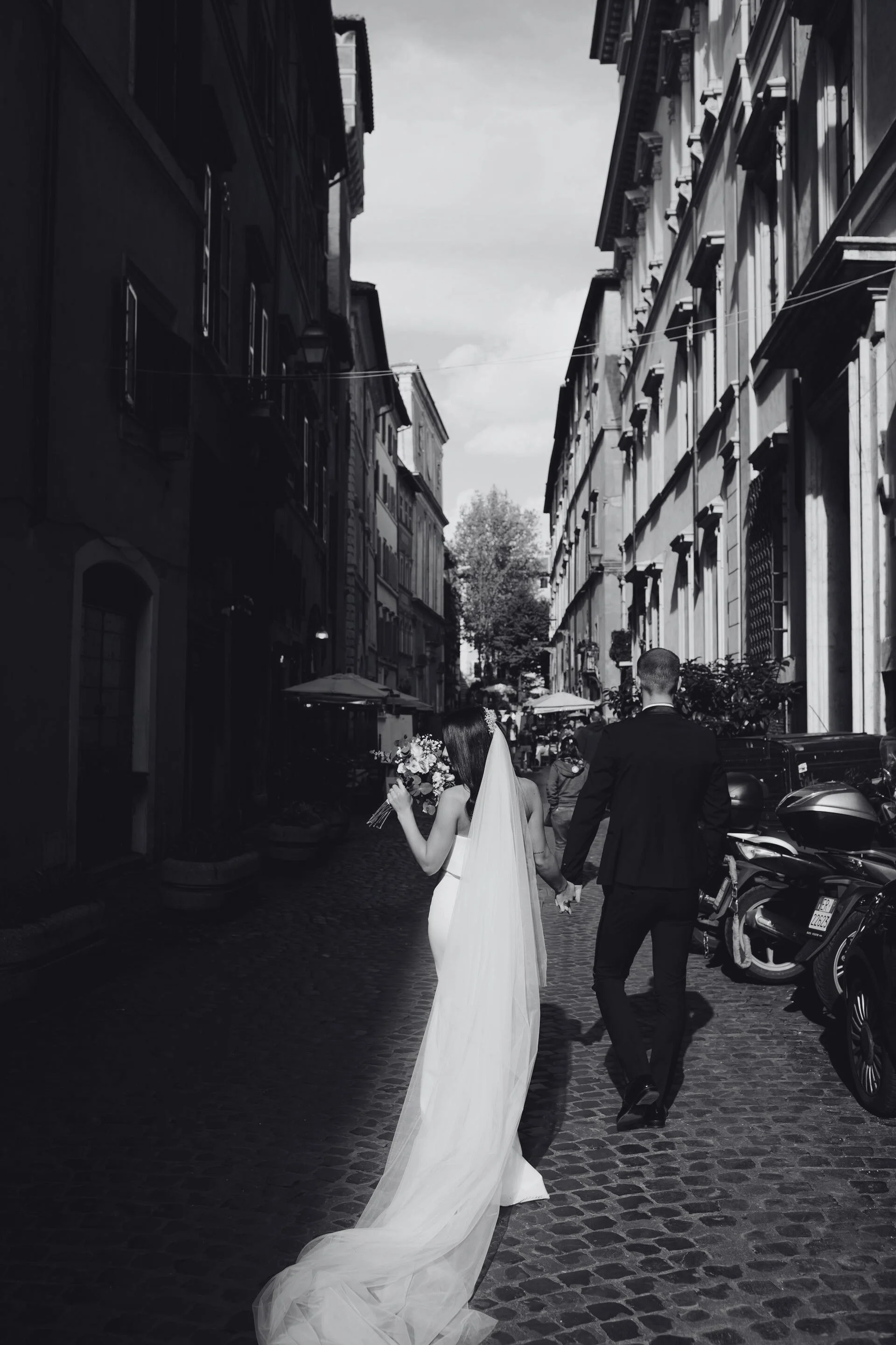 A black and white photo of a bride and groom walking hand in hand down a cobblestone street, with the bride holding a bouquet of flowers and wearing a long veil and gown. The street has tall buildings on both sides with outdoor cafes and parked motorcycles.