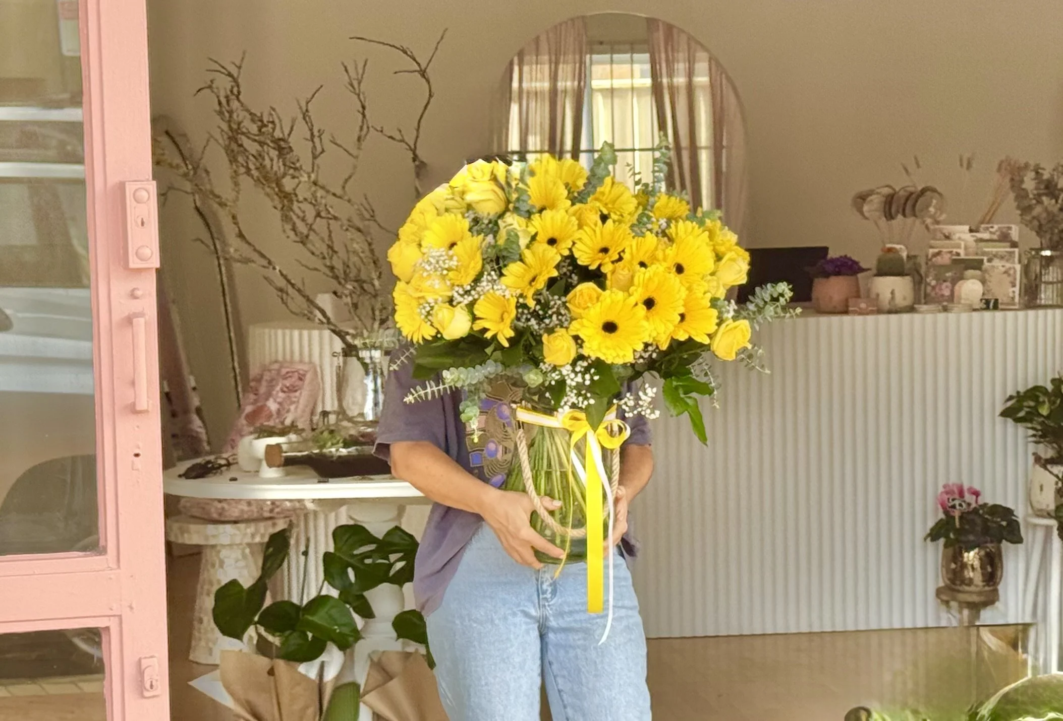 Person holding a large bouquet of yellow flowers in a floral shop or room with various plants and decorative items.