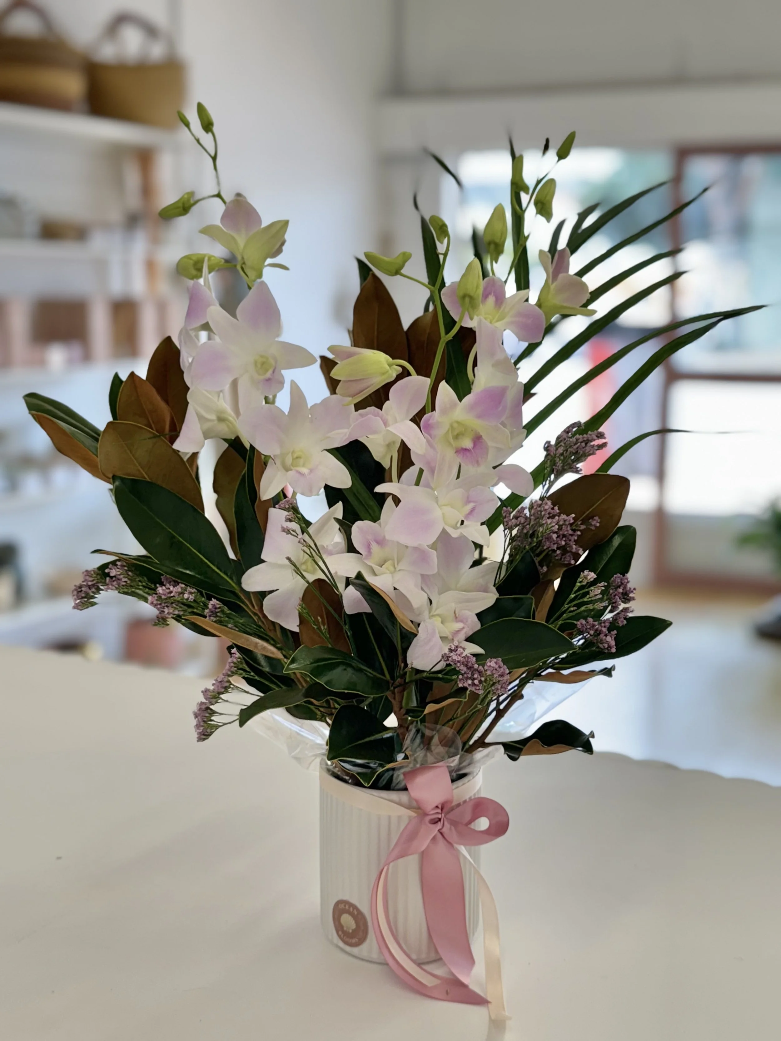 A floral arrangement with pink and white orchids, green and brown leaves, and small purple flowers in a white striped vase tied with a pink ribbon.