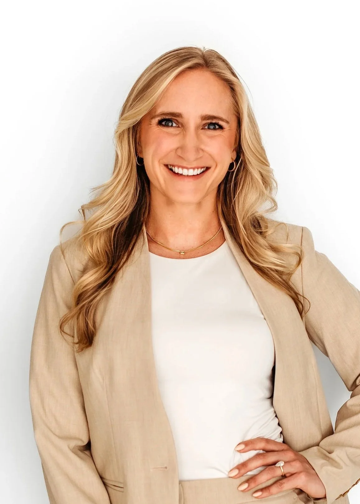 A woman with long blonde hair, smiling, wearing a beige blazer over a white top, with jewelry including earrings, a necklace, and a ring, standing against a white background.