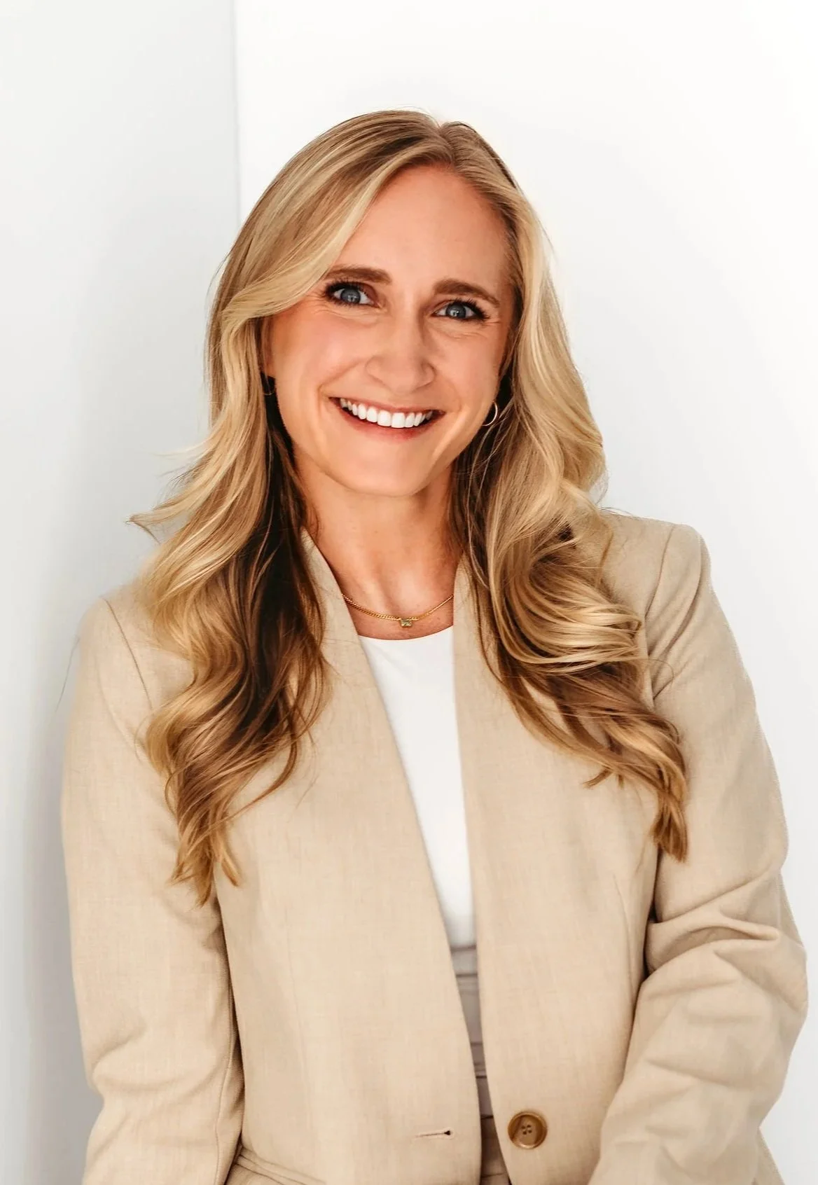 A woman with long blonde hair, smiling, wearing a beige blazer over a white top, with jewelry including earrings, a necklace, and a ring, standing against a white background.