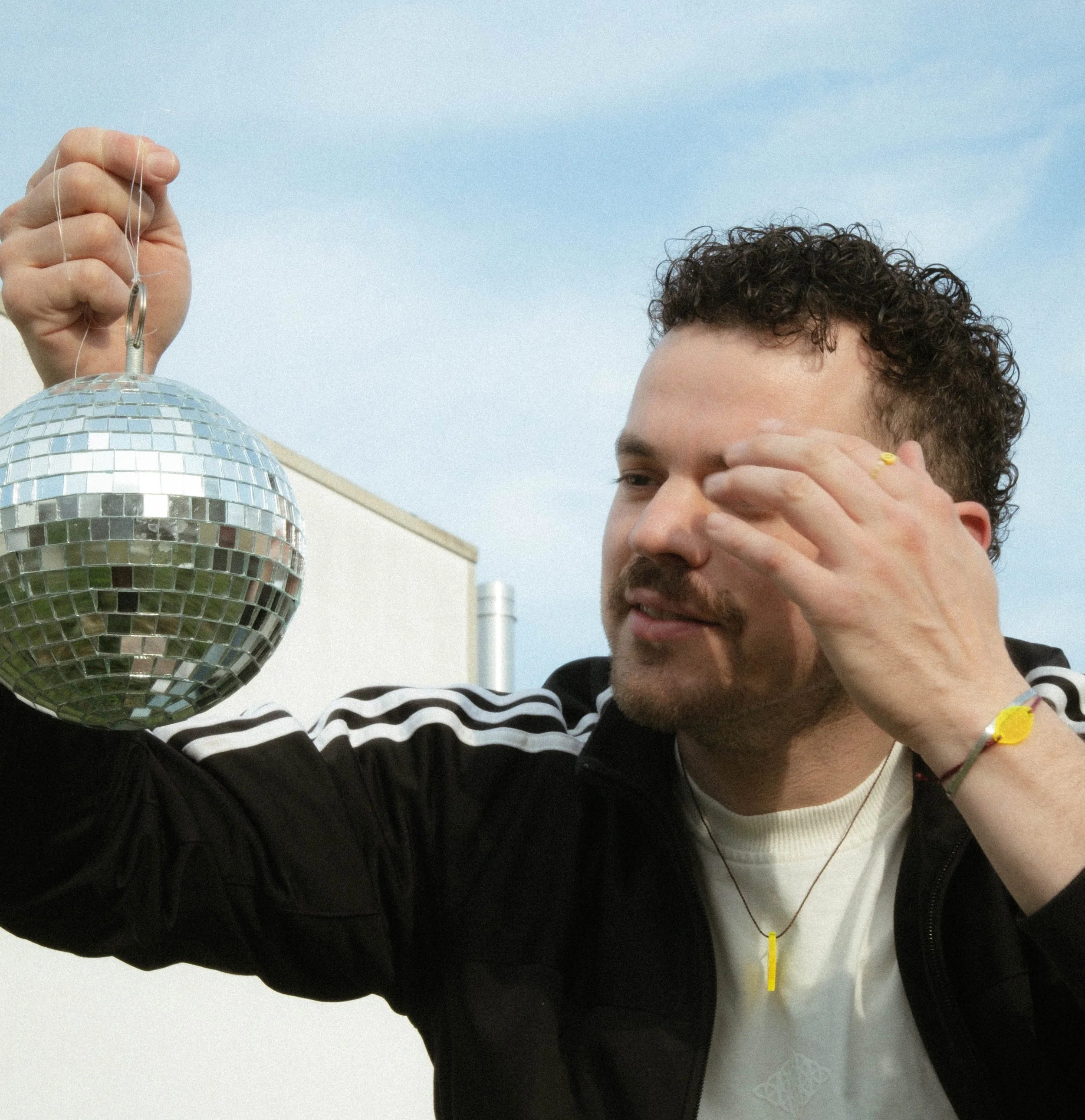 A man with curly hair, a beard, and wearing a black jacket is holding a small disco ball ornament. He is using one hand to touch his face and is outdoors against a blue sky.
