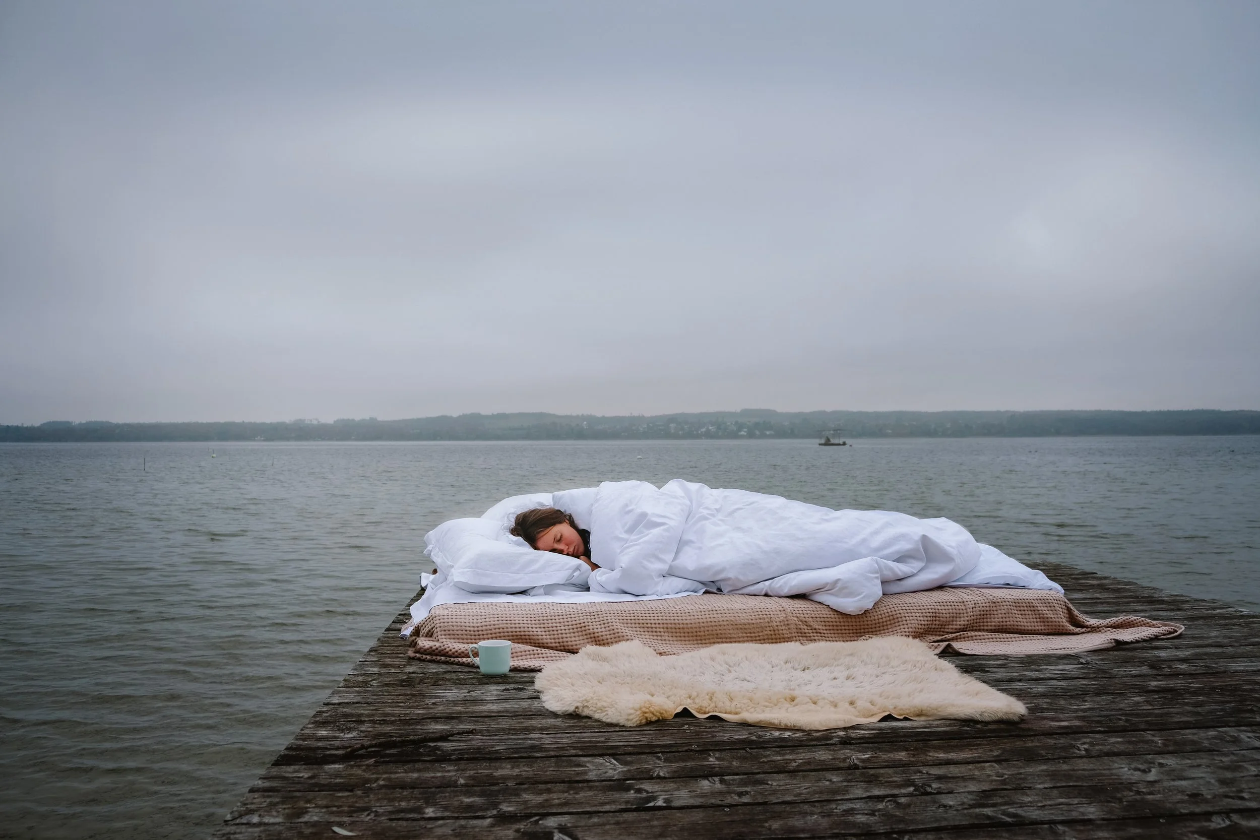 A woman sleeping on a bed with white bedding on a wooden dock by the water, with a cloudy sky overhead.