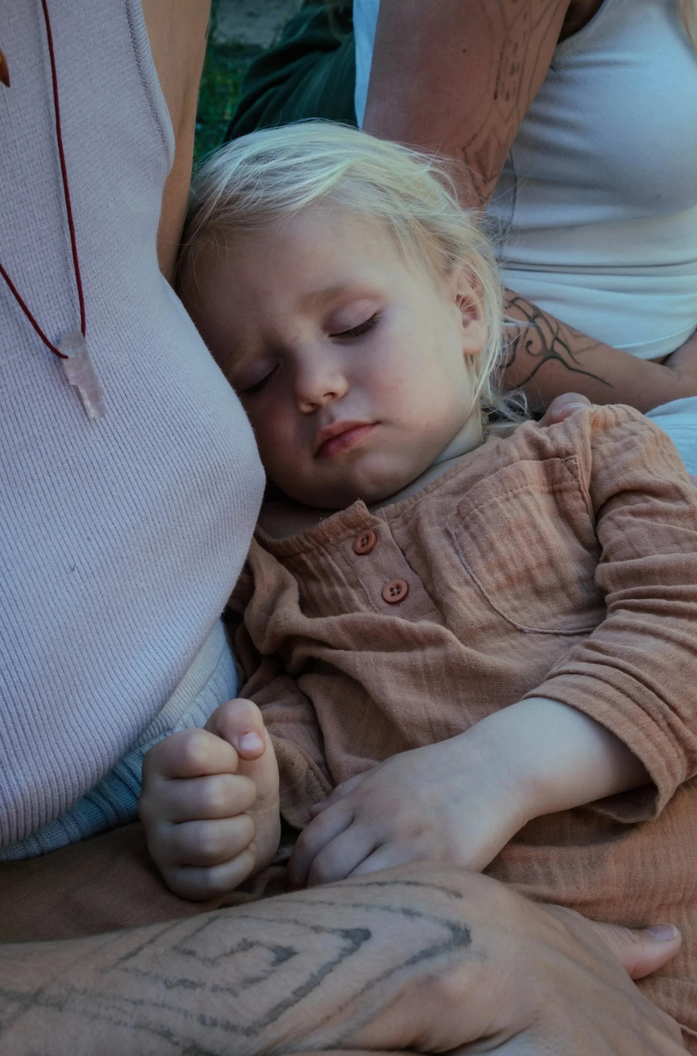 A young girl with blonde hair and closed eyes is sleeping with her head resting on an adult's chest, who is wearing a beige tank top. The girl is wearing a rust-colored shirt with buttons and has her hand clenched into a loose fist.