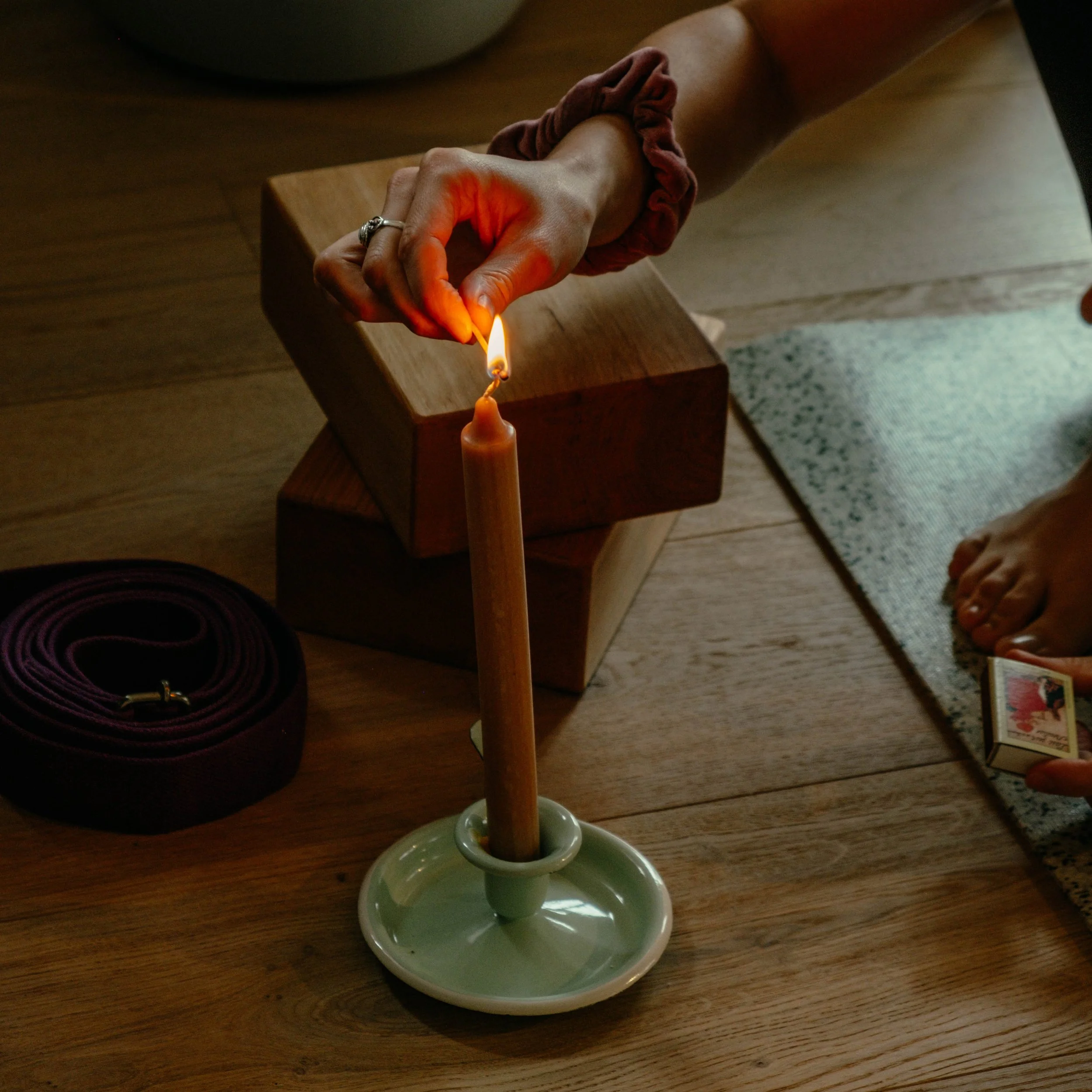 Person lighting a candle on a wooden table with a matchstick, next to a rolled-up belt and a small box, with feet visible on a small rug.