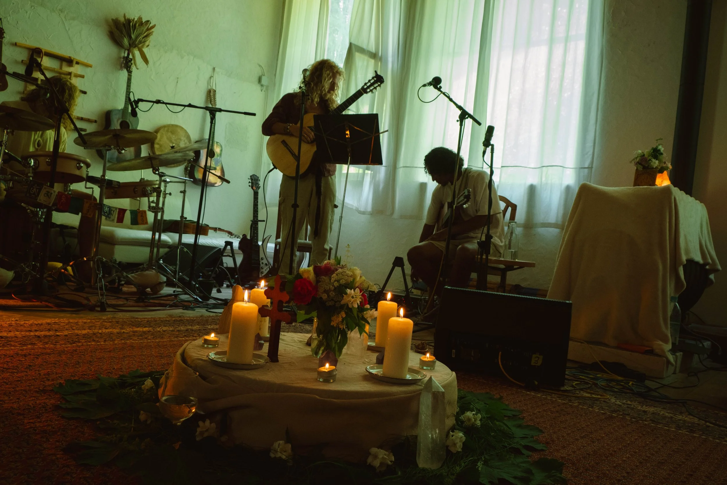 Musicians performing in a softly lit indoor setting with candles, flowers, and a religious cross in the foreground.
